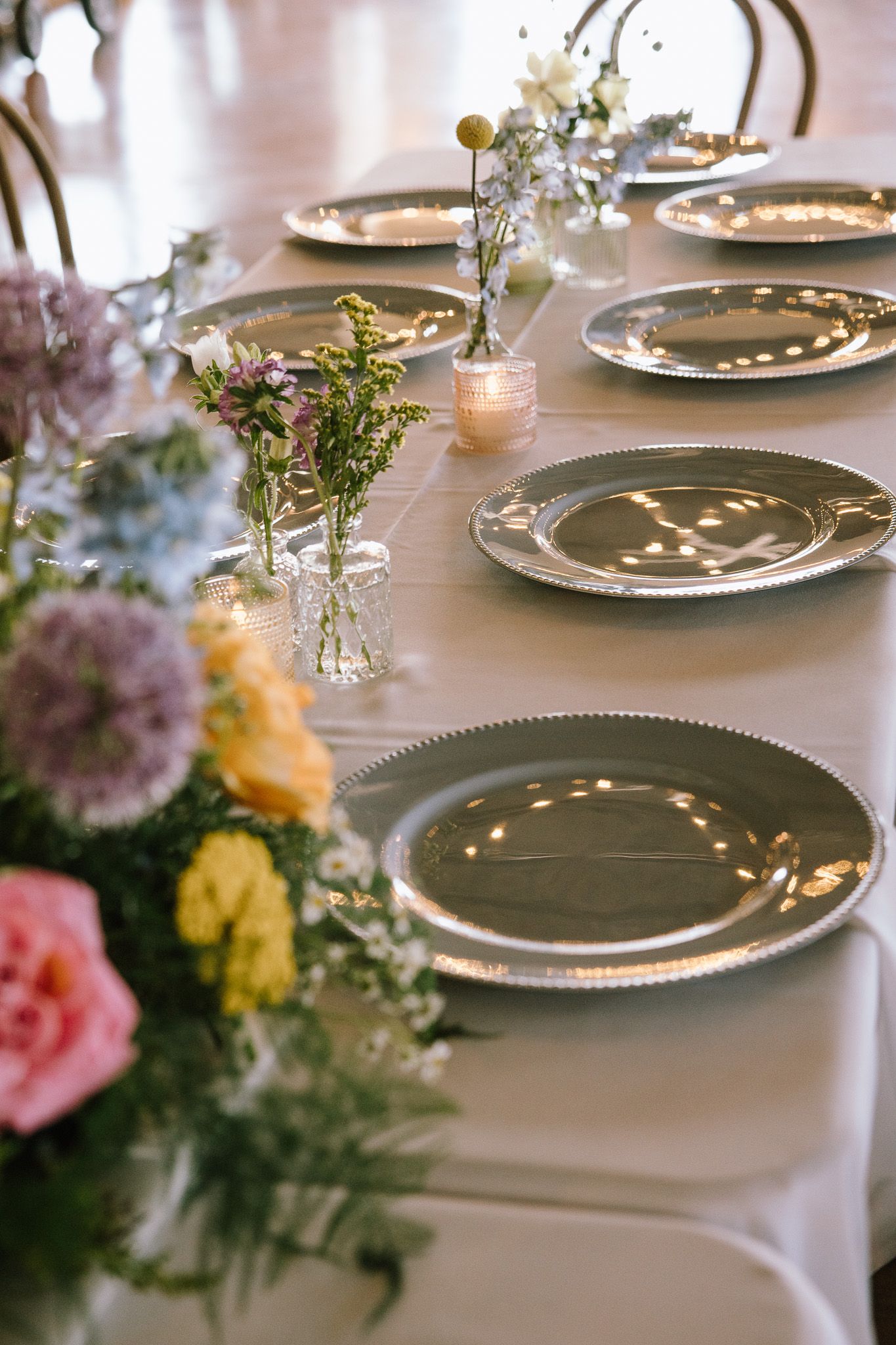 Elegant wedding table setting with silver charger plates, pastel floral arrangements, and candles on a white linen tablecloth.
