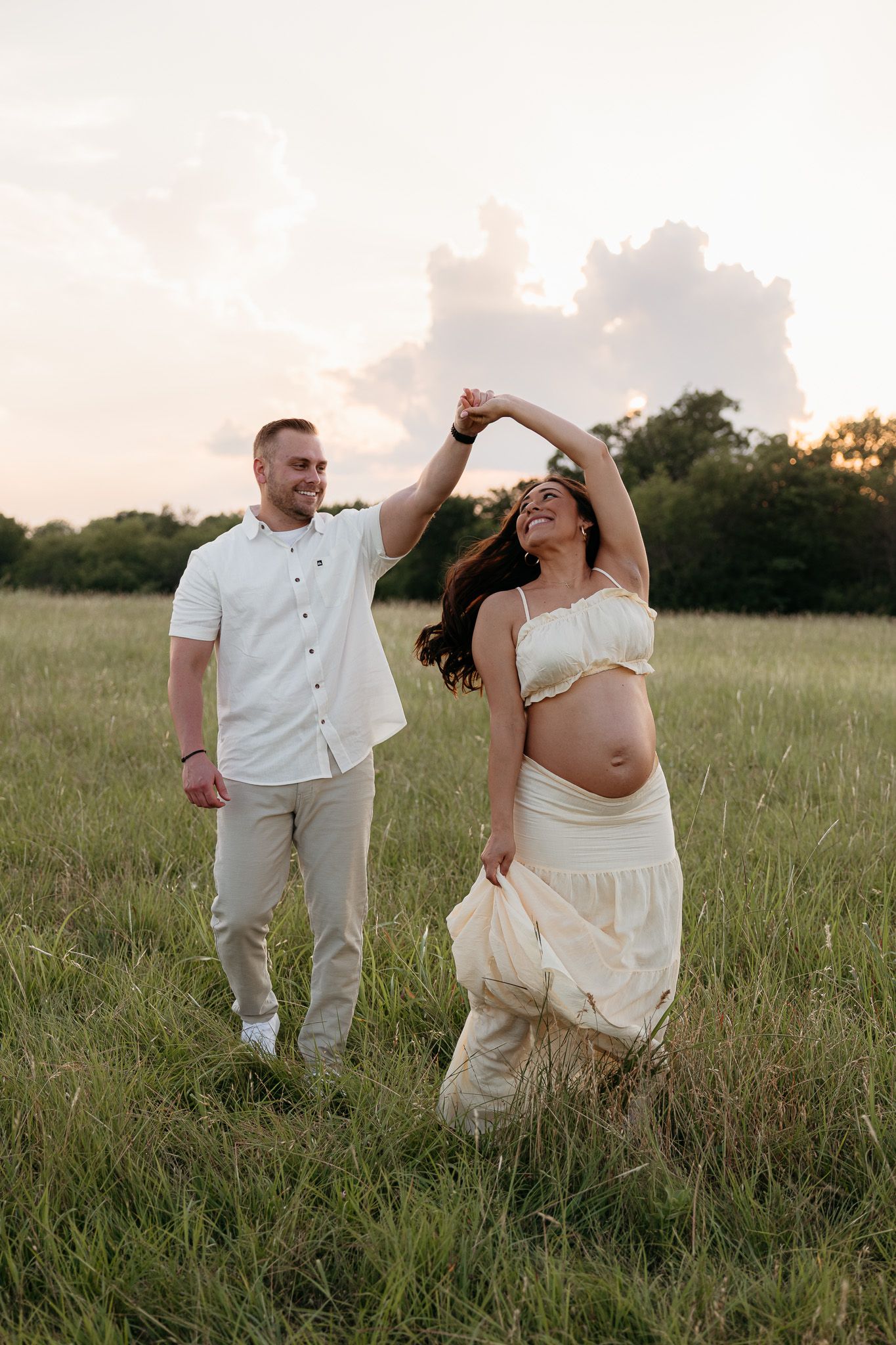 A happy couple dances in a green field at sunset, the smiling pregnant woman twirling in a cream dress.