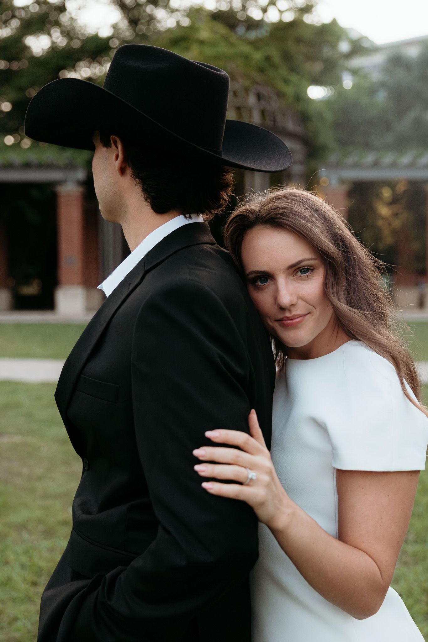 Bride in white dress embraces groom in black suit and cowboy hat outdoors at elegant wedding venue.