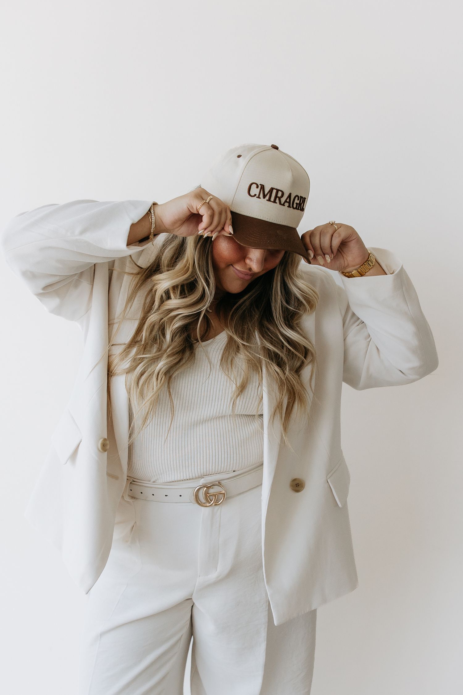 Woman in white blazer and pants wearing a beige CMRAGE snapback cap, posing against a white background.