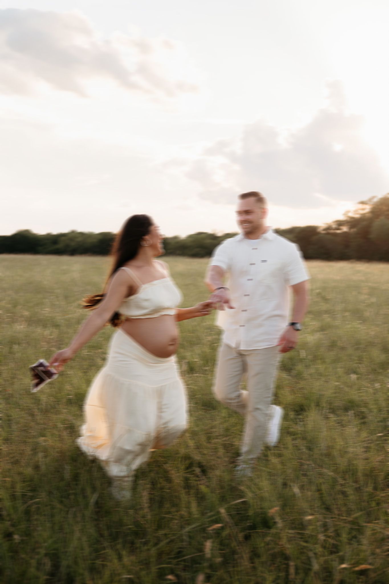 Pregnant woman in white dress holds hands with man running through green field during golden hour maternity photoshoot.