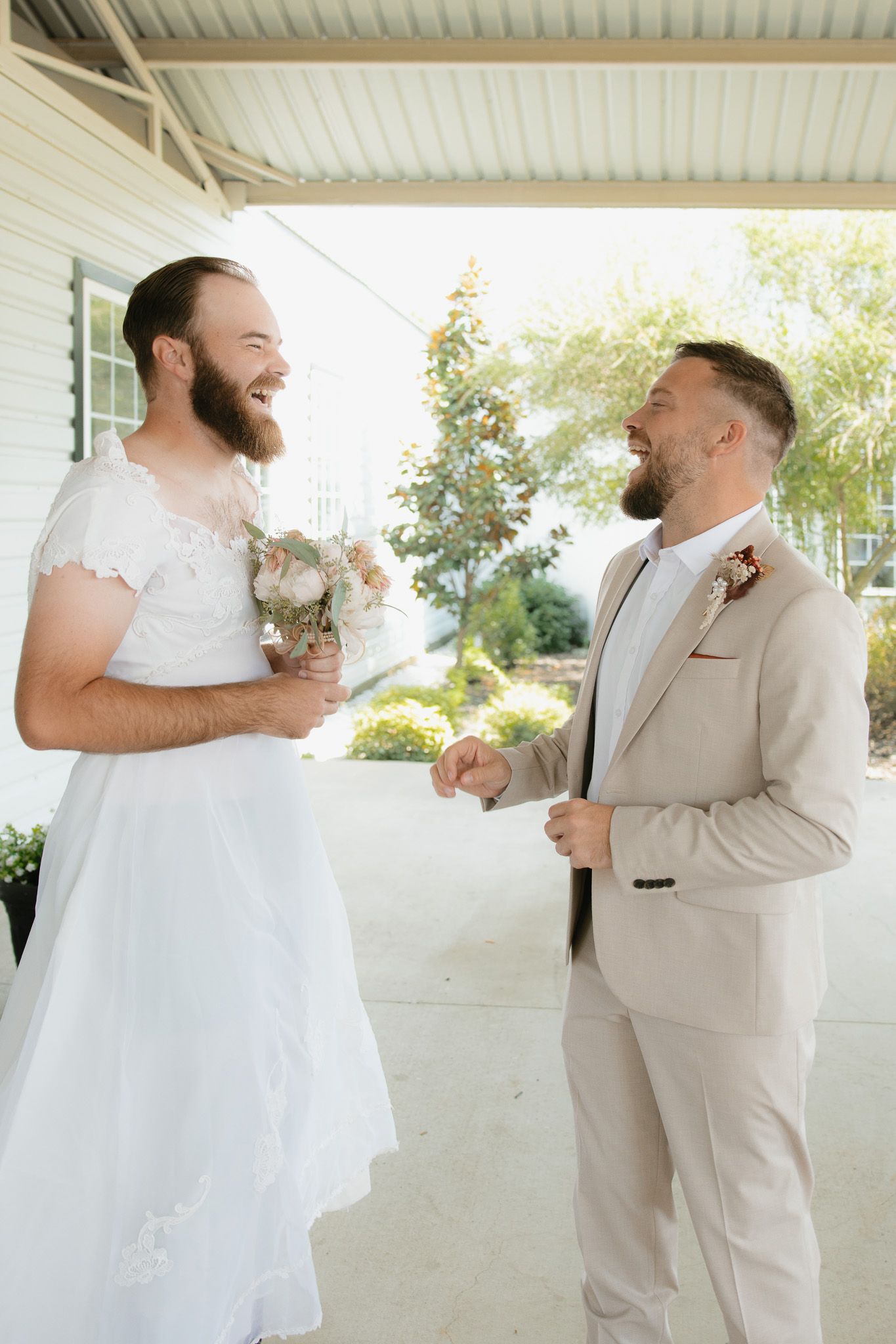 Two grooms in wedding attire share a joyful moment, one wearing a white dress holding flowers, the other in a beige suit.