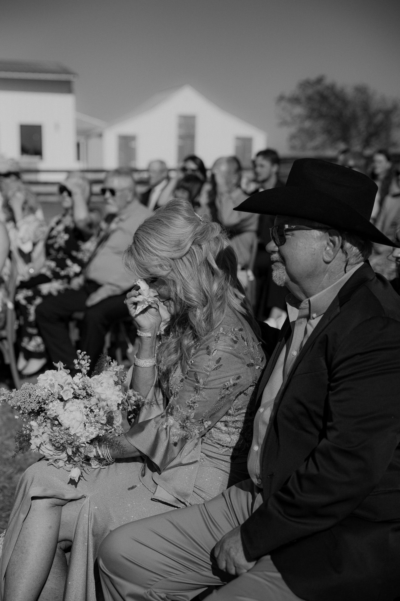 Black and white photo of emotional bride wiping tears holding bouquet seated beside man in cowboy hat at outdoor wedding ceremony.