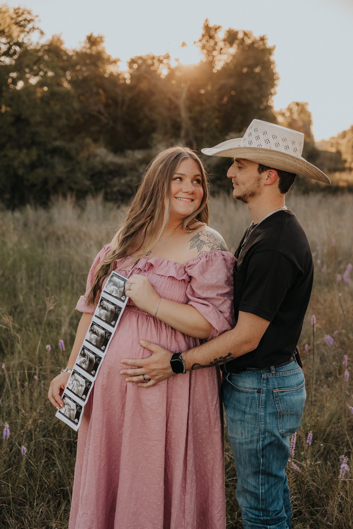 Couple in rustic attire sharing tender moment in wildflower field at sunset.