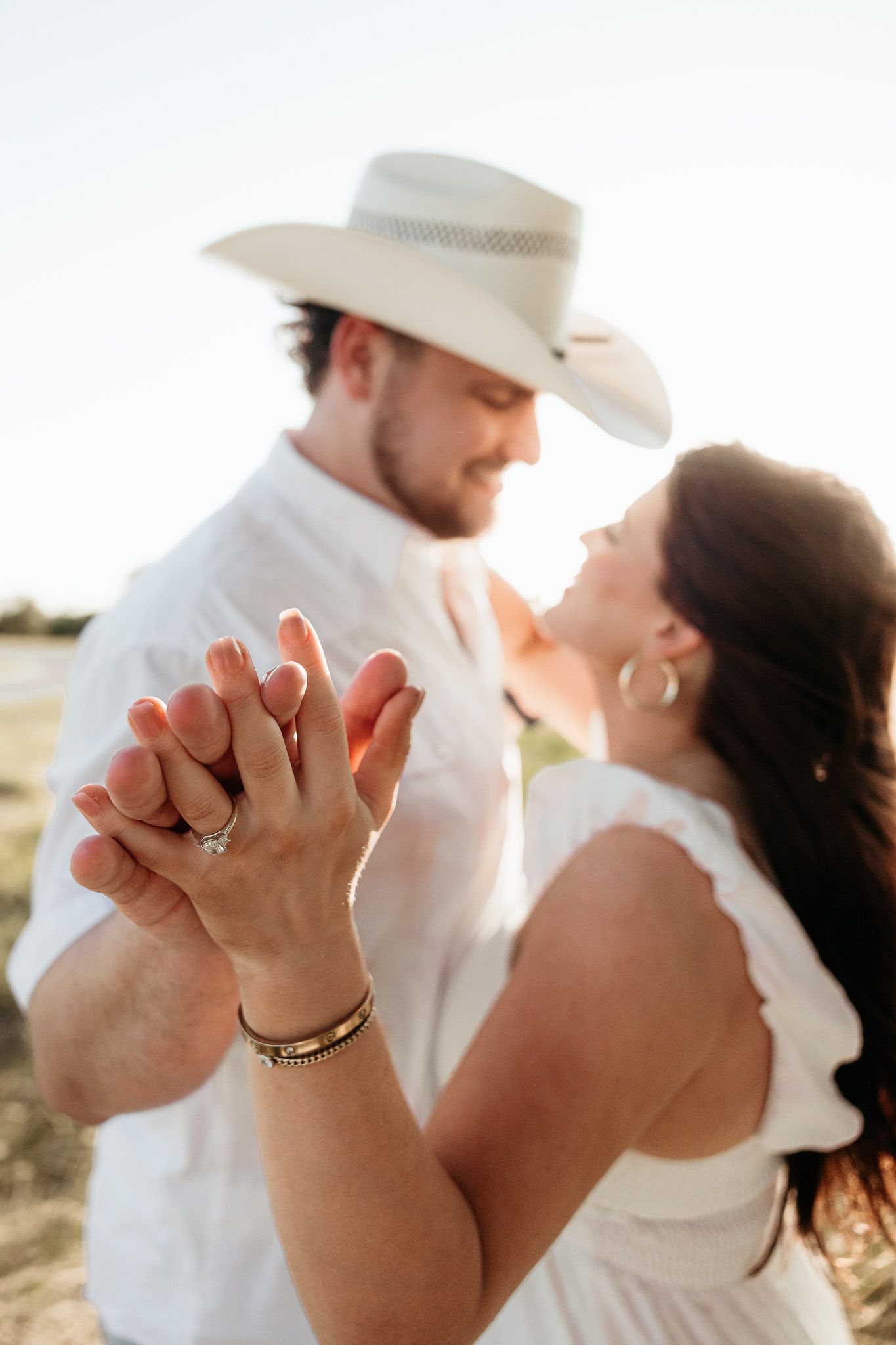 Couple in white outfits holding hands outdoors, man wearing cowboy hat, woman showing engagement ring, golden hour lighting.