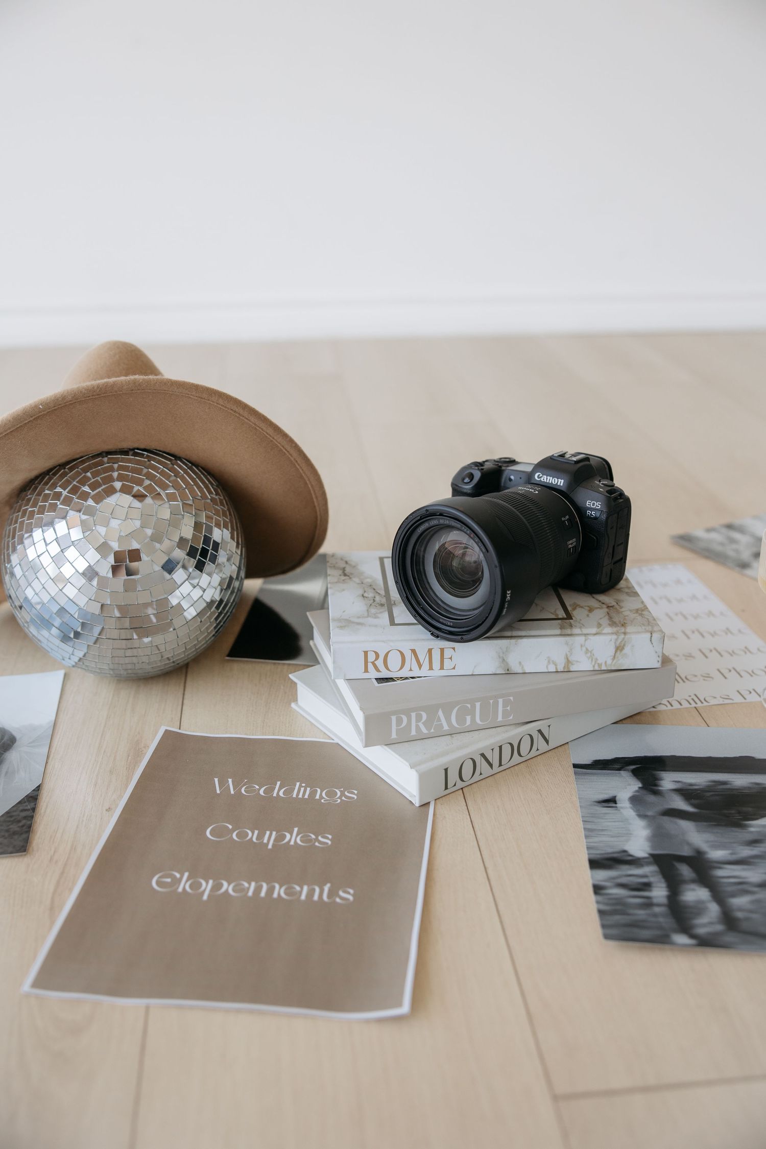 Canon camera on travel books beside a disco ball, tan hat, and wedding photography brochure on wood floor.