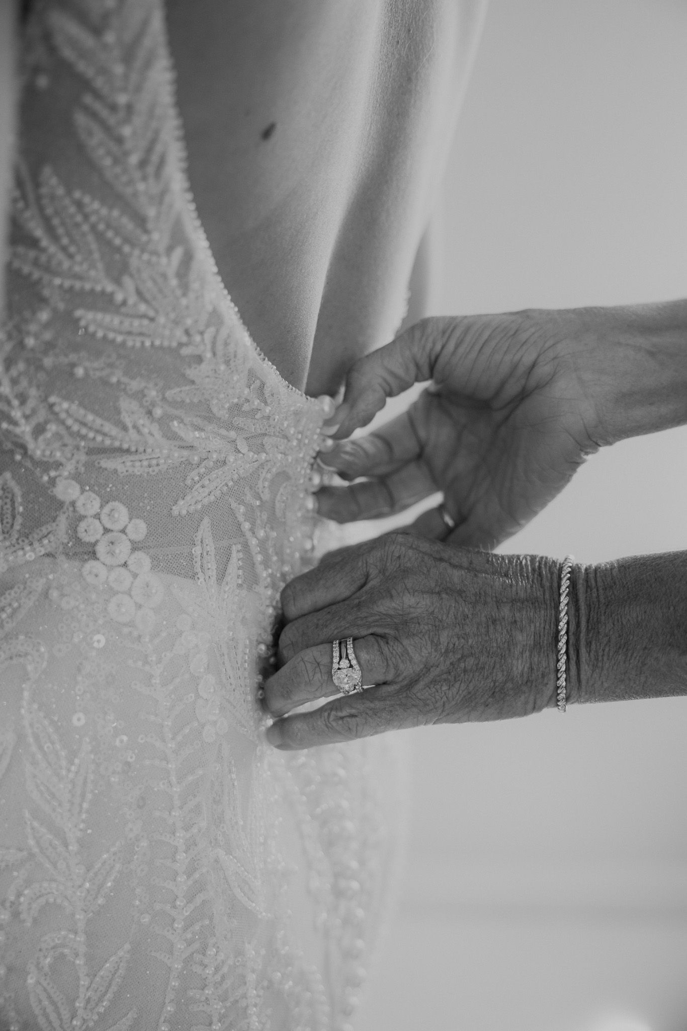 Black and white photo of elderly hands with ring and bracelet buttoning the back of a beaded lace wedding dress.
