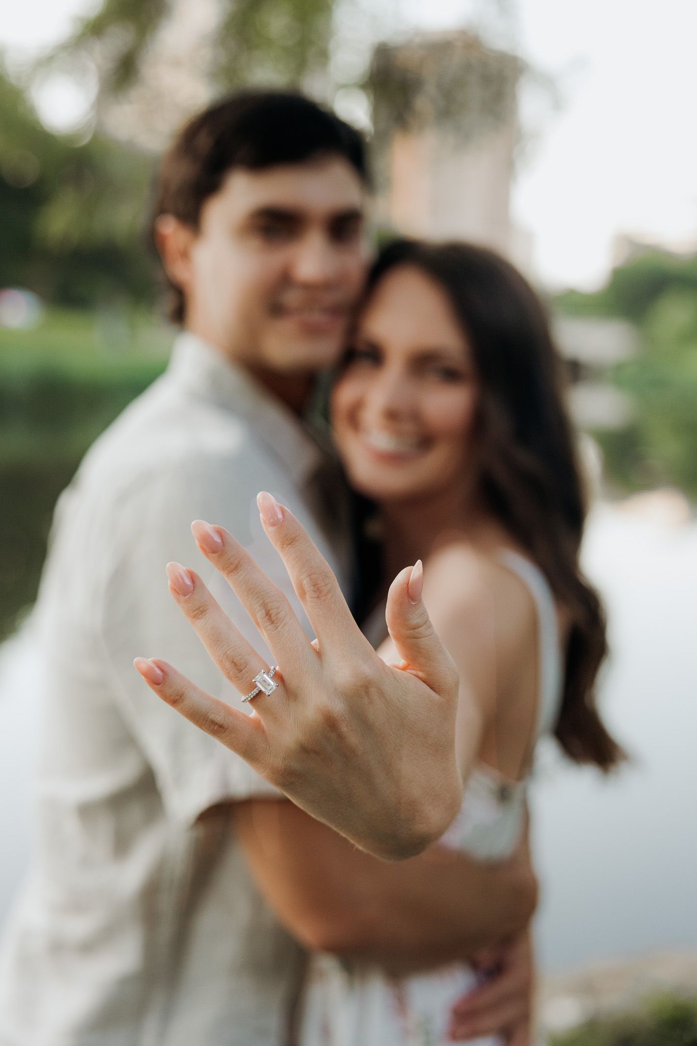 Happy couple showing off engagement ring outdoors near a lake, woman extending hand with diamond ring toward camera.
