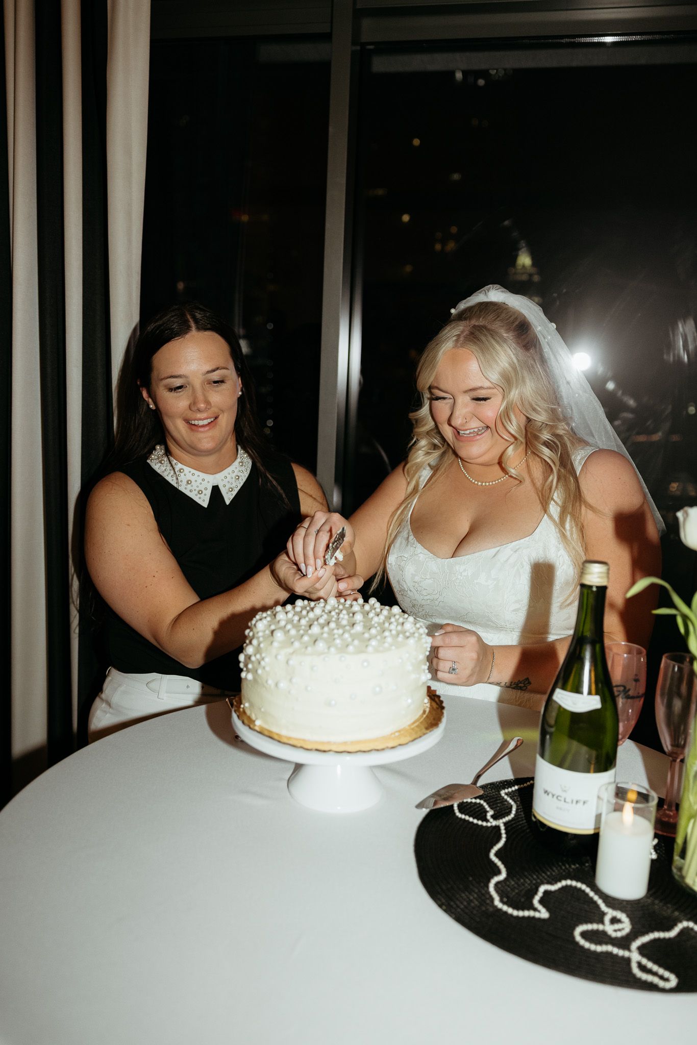 Two brides cutting a white pearl-decorated wedding cake beside a champagne bottle at an elegant evening reception.