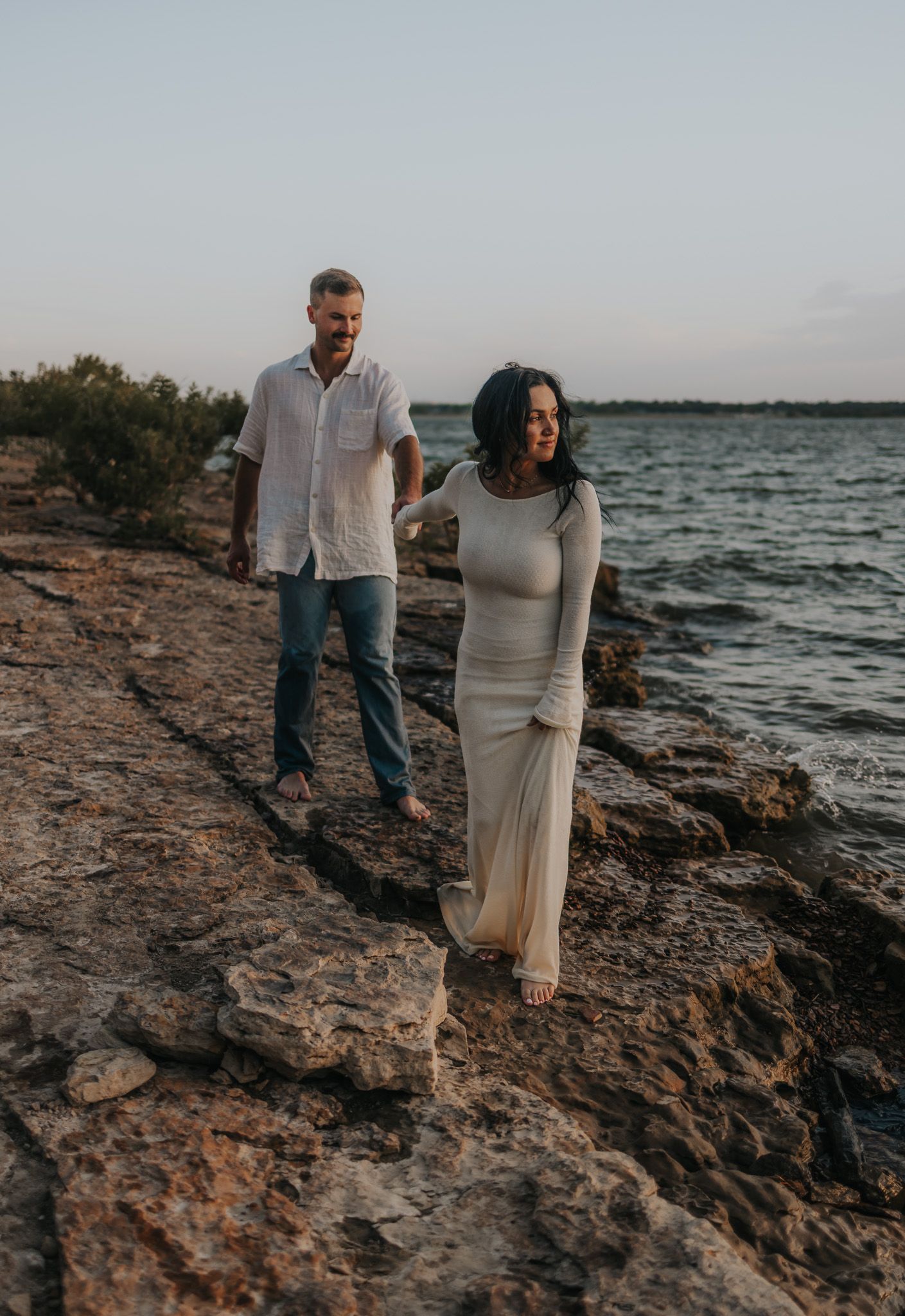 A couple stands together on rocky coastal shoreline at sunset with ocean waves in the background.
