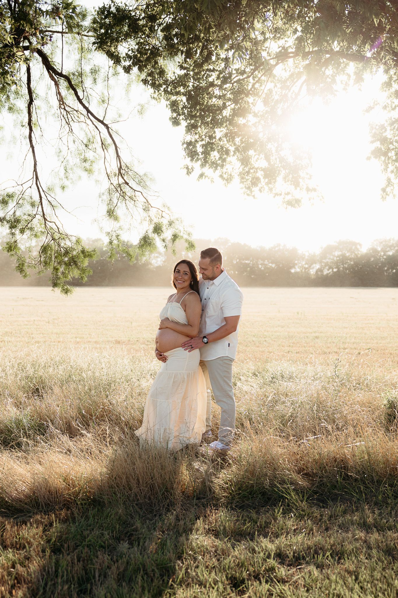 Couple poses in golden field at sunset, pregnant woman in white dress embraced by partner under tree branches.