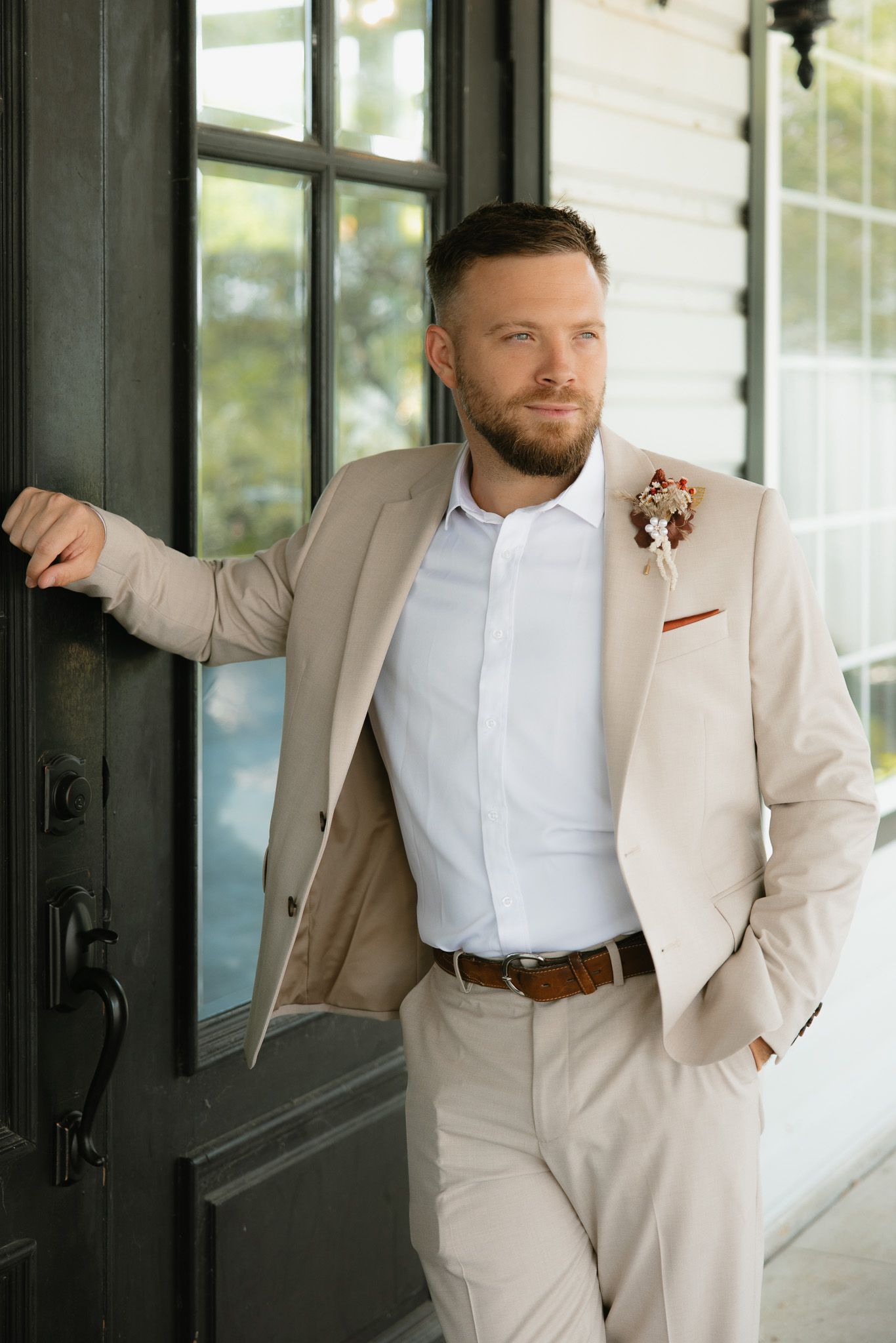 Man in beige suit and white shirt standing by black door with hand in pocket, wearing brown belt and pocket square.