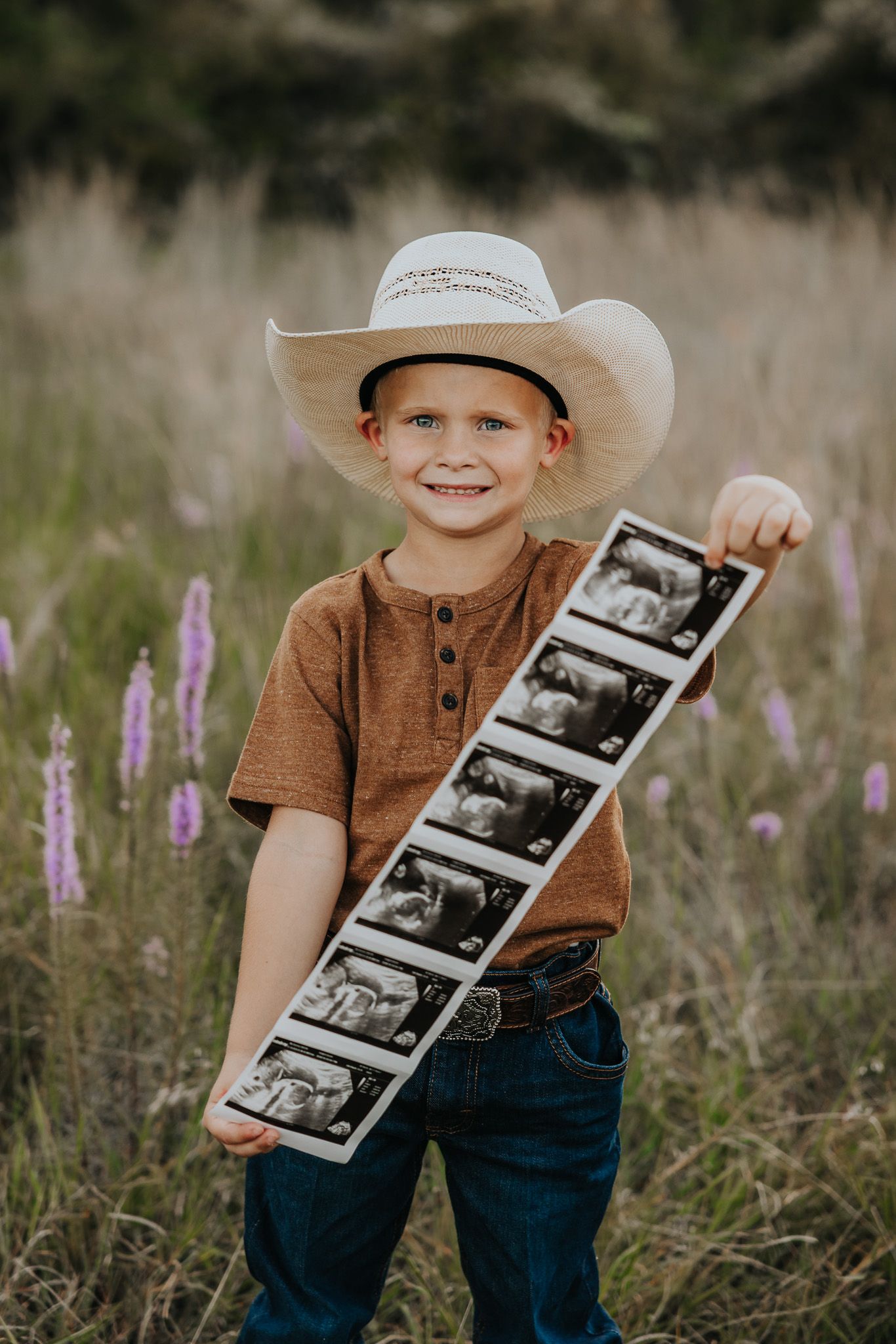 A child in a cowboy hat and brown shirt holds ultrasound photos in a field of purple wildflowers.