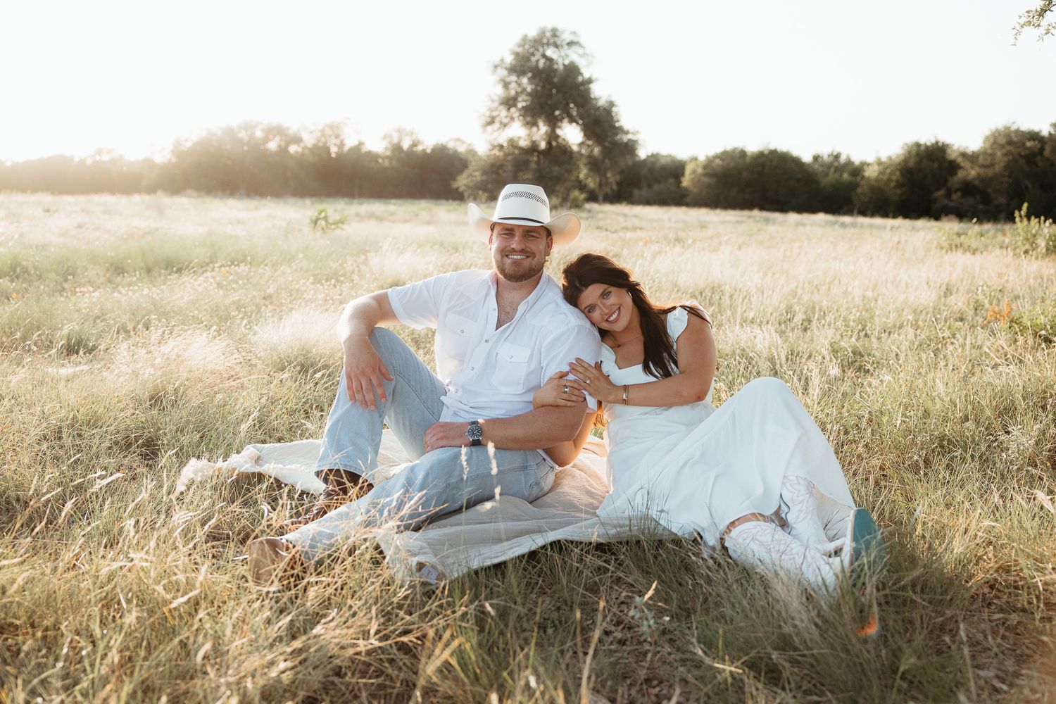 Couple sitting together in a sunlit golden field, wearing white outfits, smiling and relaxing on a blanket outdoors.