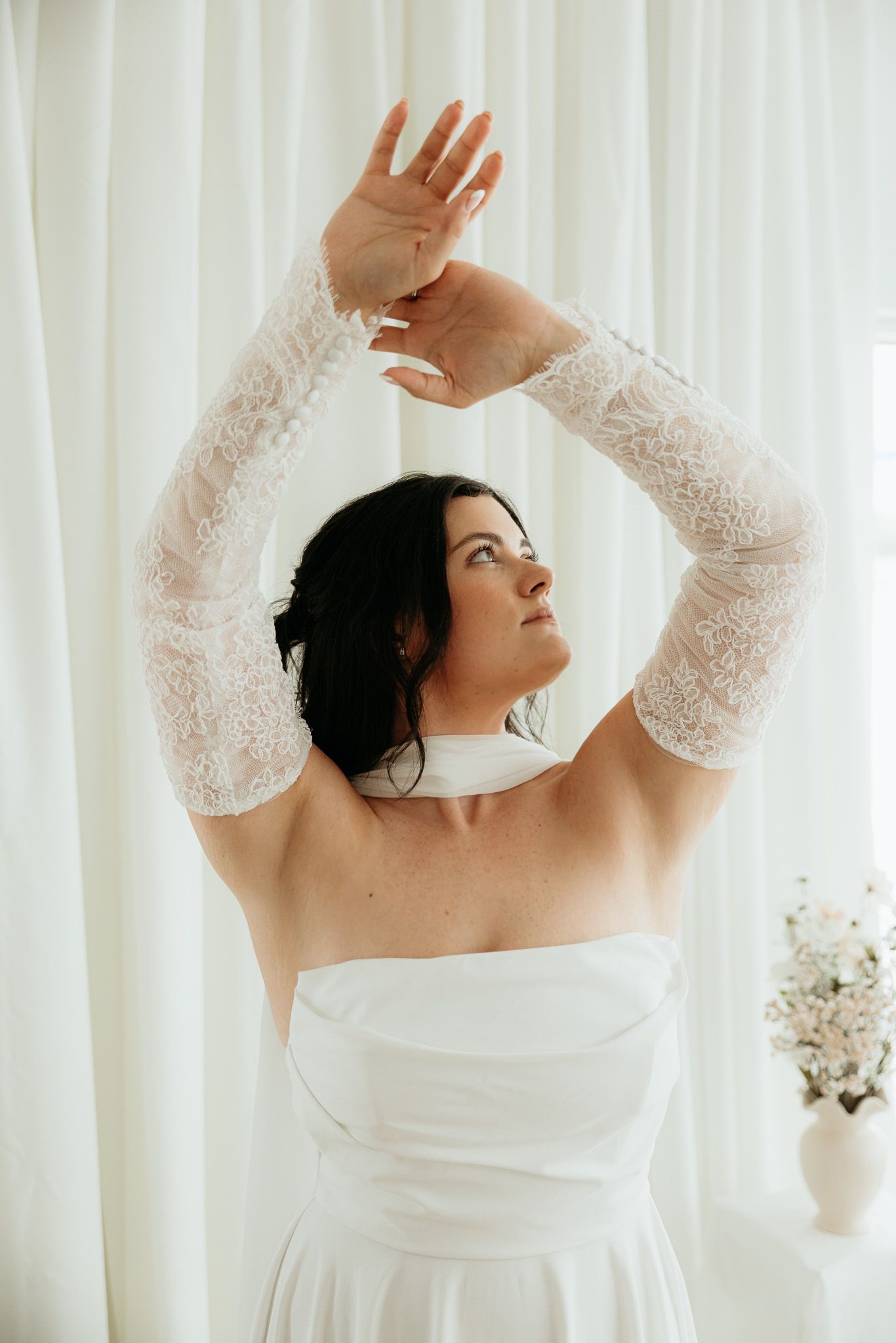 Bride in strapless white gown with lace sleeves raises arms gracefully against sheer white curtains.