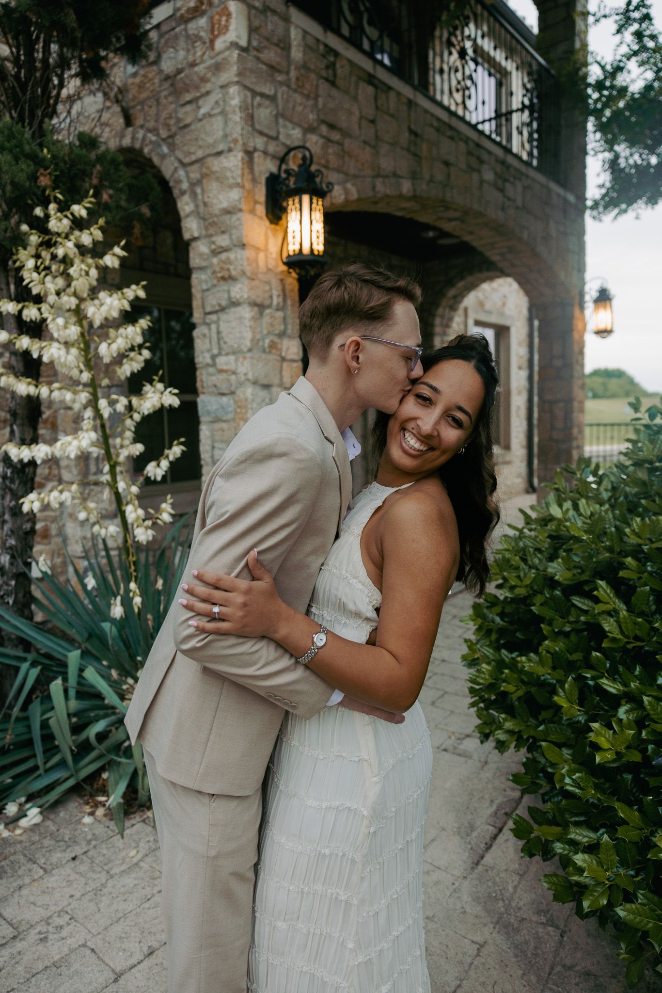 A couple embraces outside a stone building, the groom kissing his bride on the cheek as she smiles joyfully.