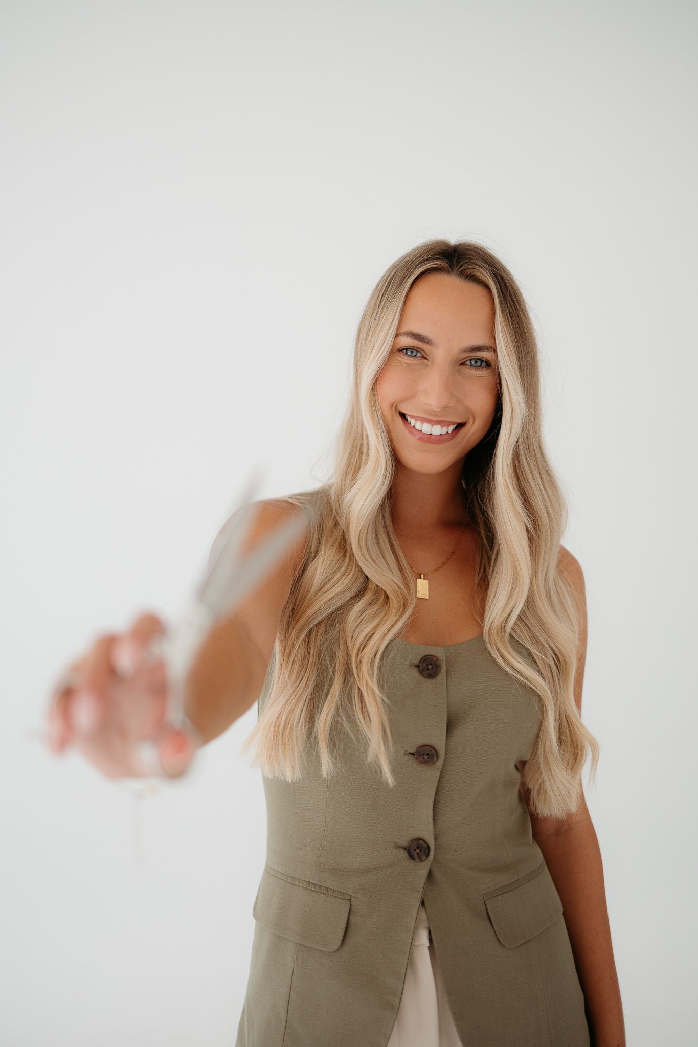Smiling blonde woman in olive green vest holding a glass of wine toward camera against white background.