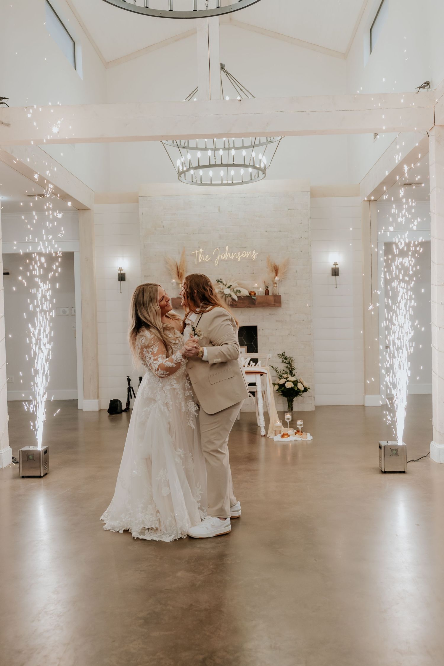 Two brides share their first dance surrounded by sparkling cold sparklers in an elegant white wedding venue.