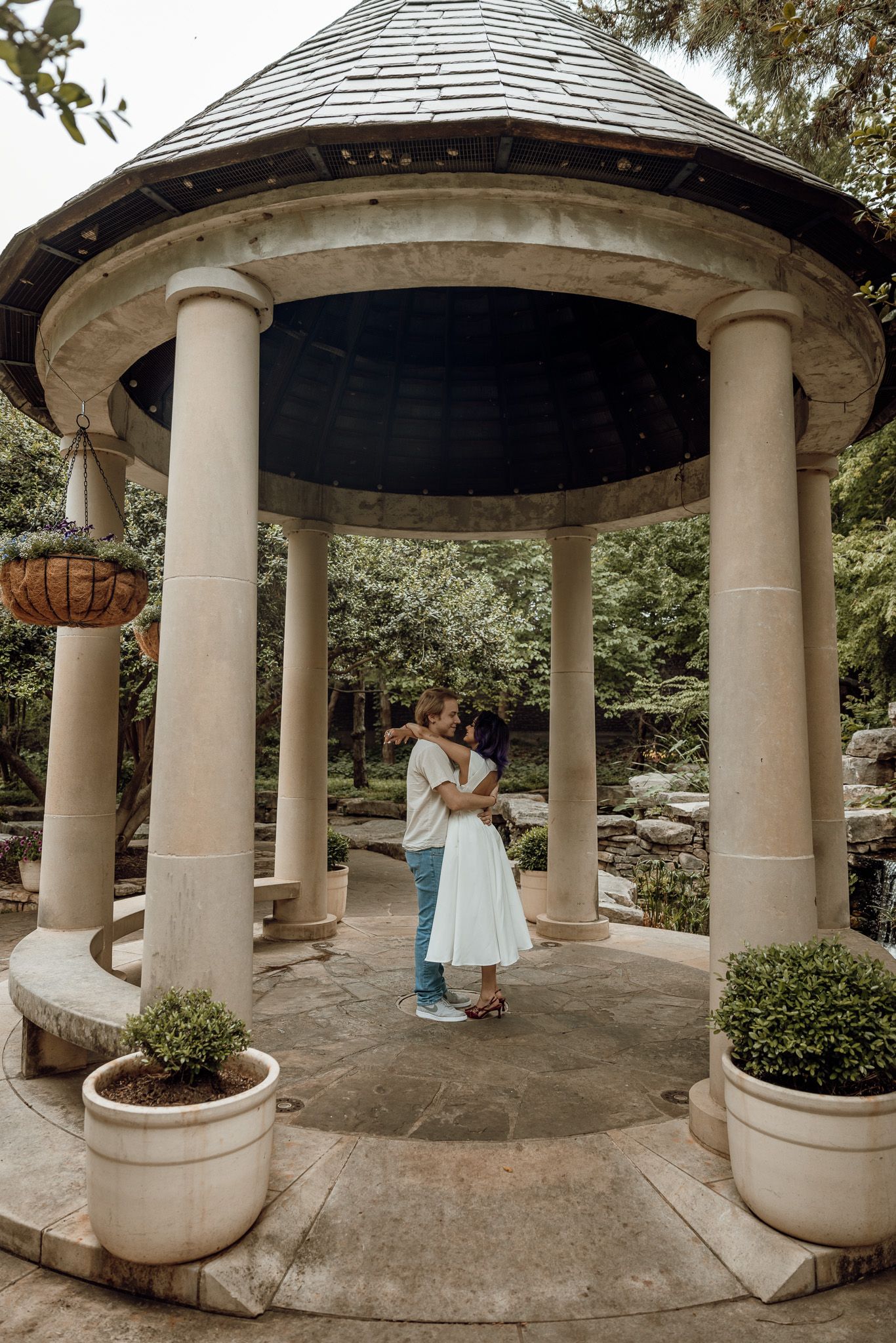 Couple embracing romantically under an elegant stone gazebo with columns surrounded by lush greenery in a garden setting.