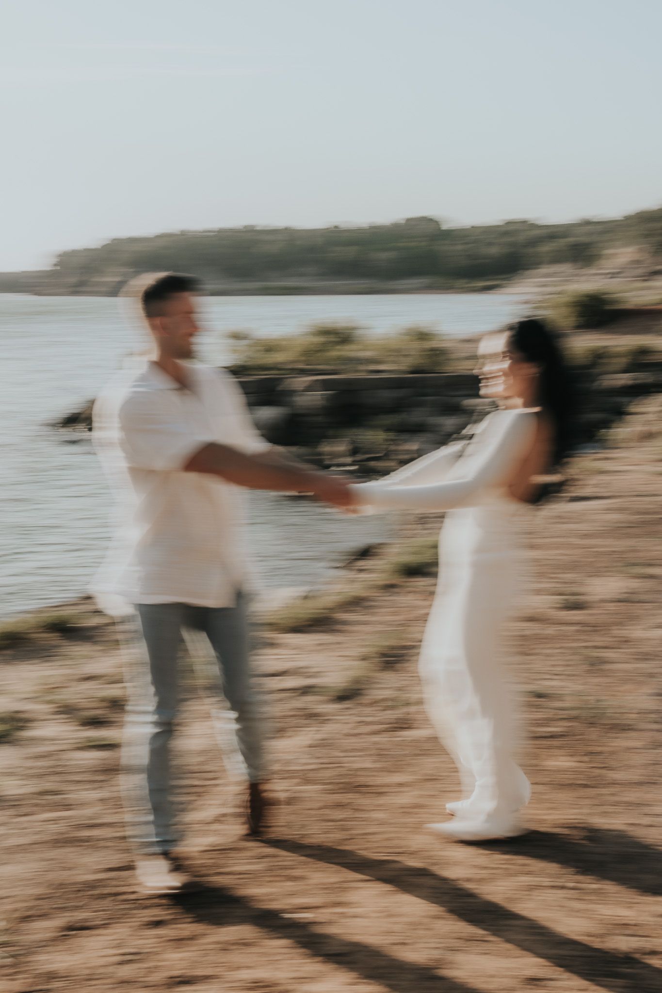 A couple in white outfits holding hands on a rocky beach at sunset, creating a dreamy, romantic atmosphere.