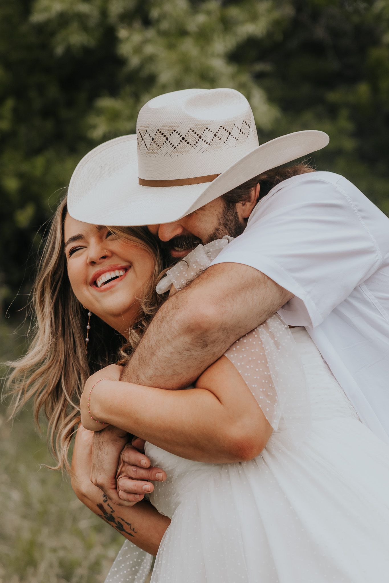 A couple embraces outdoors in casual white outfits, with one wearing a western-style cowboy hat in a candid moment.