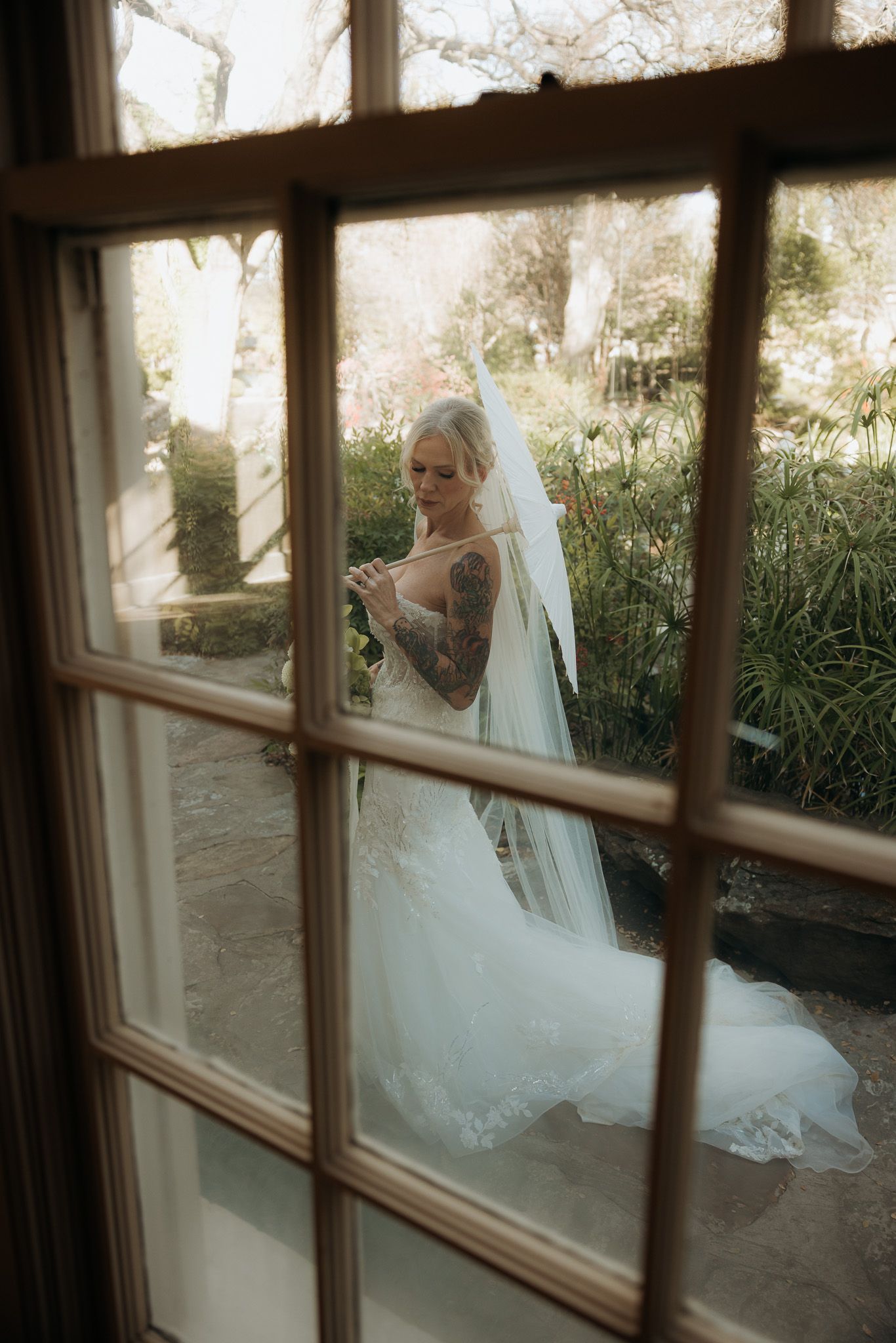 View through window panes of a bride in white dress and veil looking at phone in garden setting.