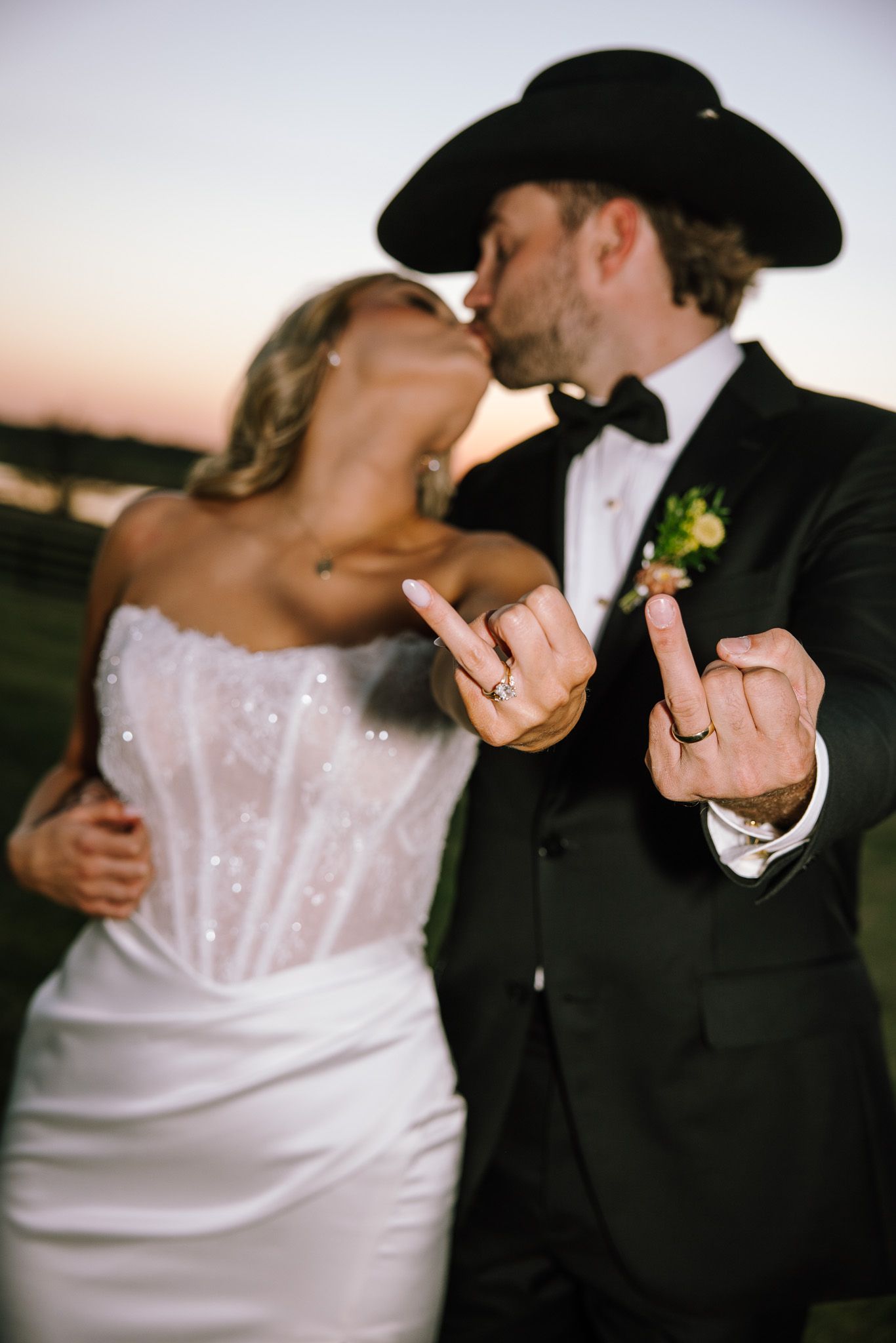Newlyweds kissing at sunset, showing off wedding rings, groom in black tuxedo and cowboy hat, bride in strapless white gown.