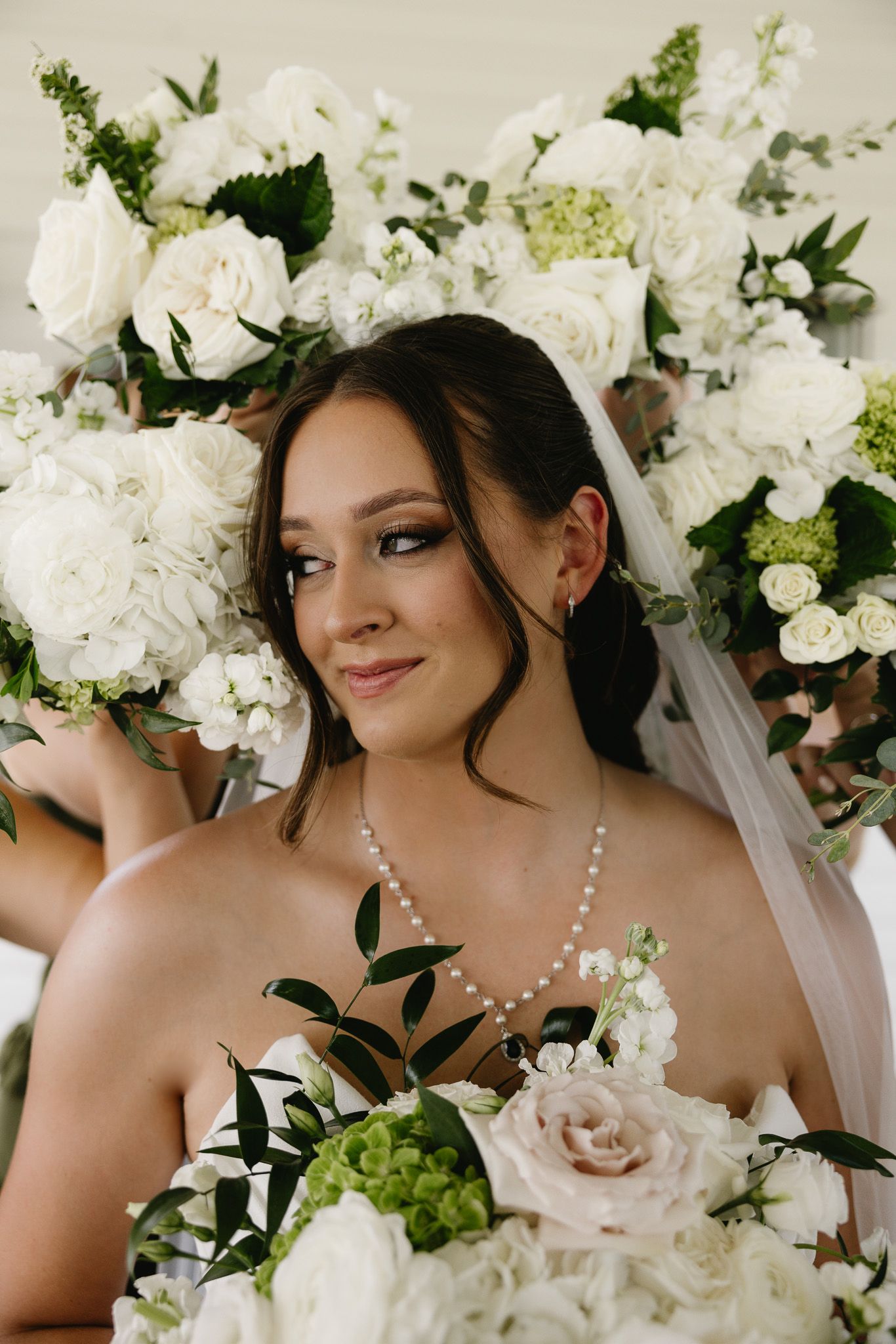 Bride in pearl necklace and veil holds white floral bouquet surrounded by elegant white roses and greenery.