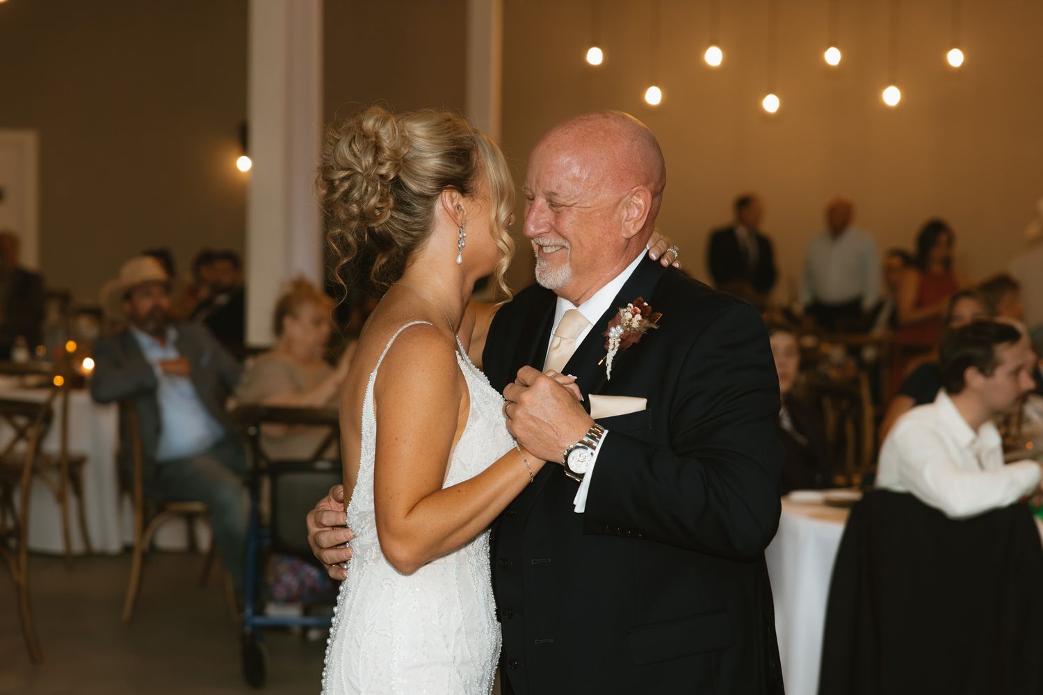 Bride and father share an emotional first dance at wedding reception with string lights and guests in background.
