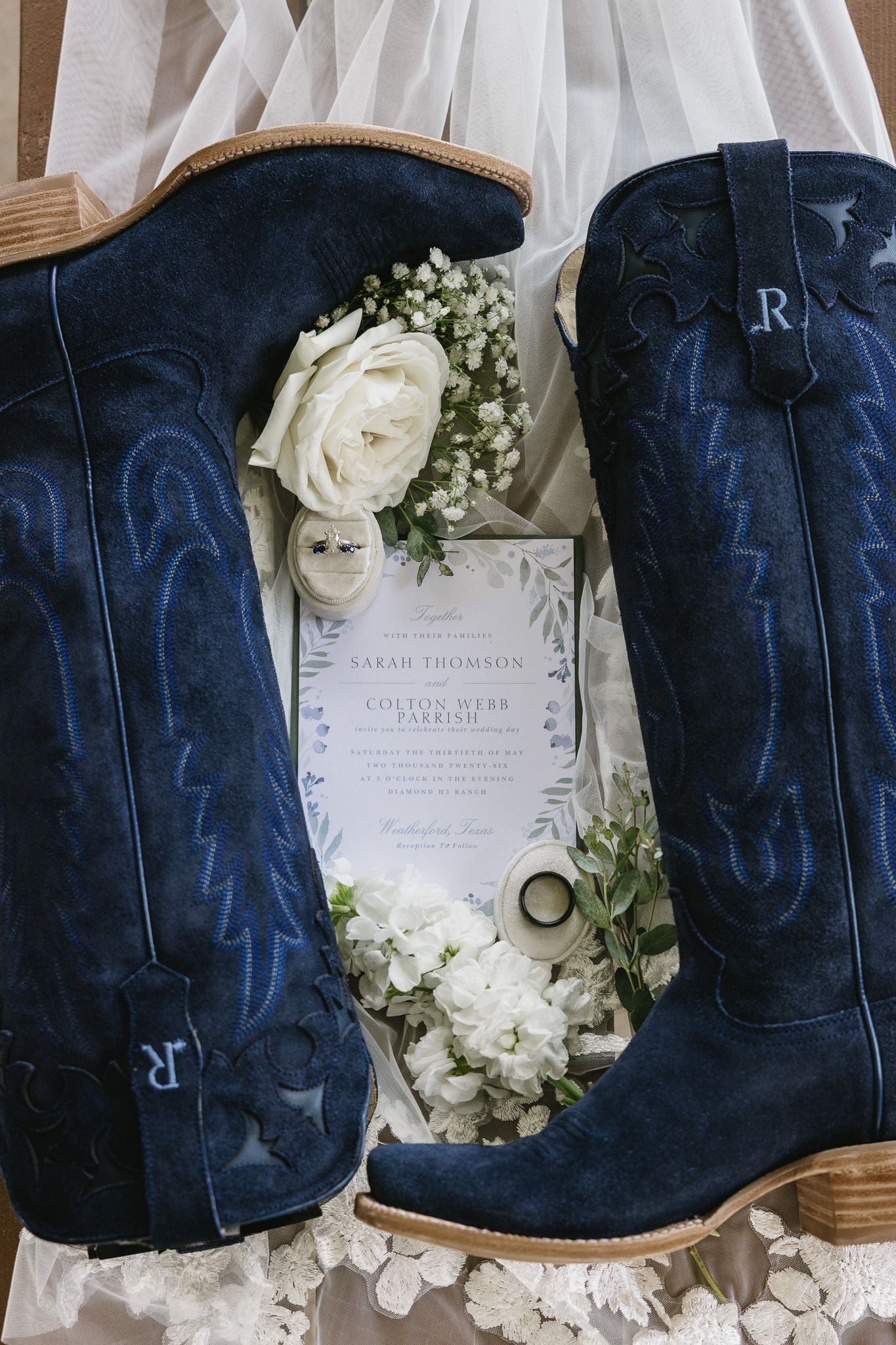Blue suede cowboy boots with wedding invitation, white roses, and rings on a bridal veil for a country wedding.