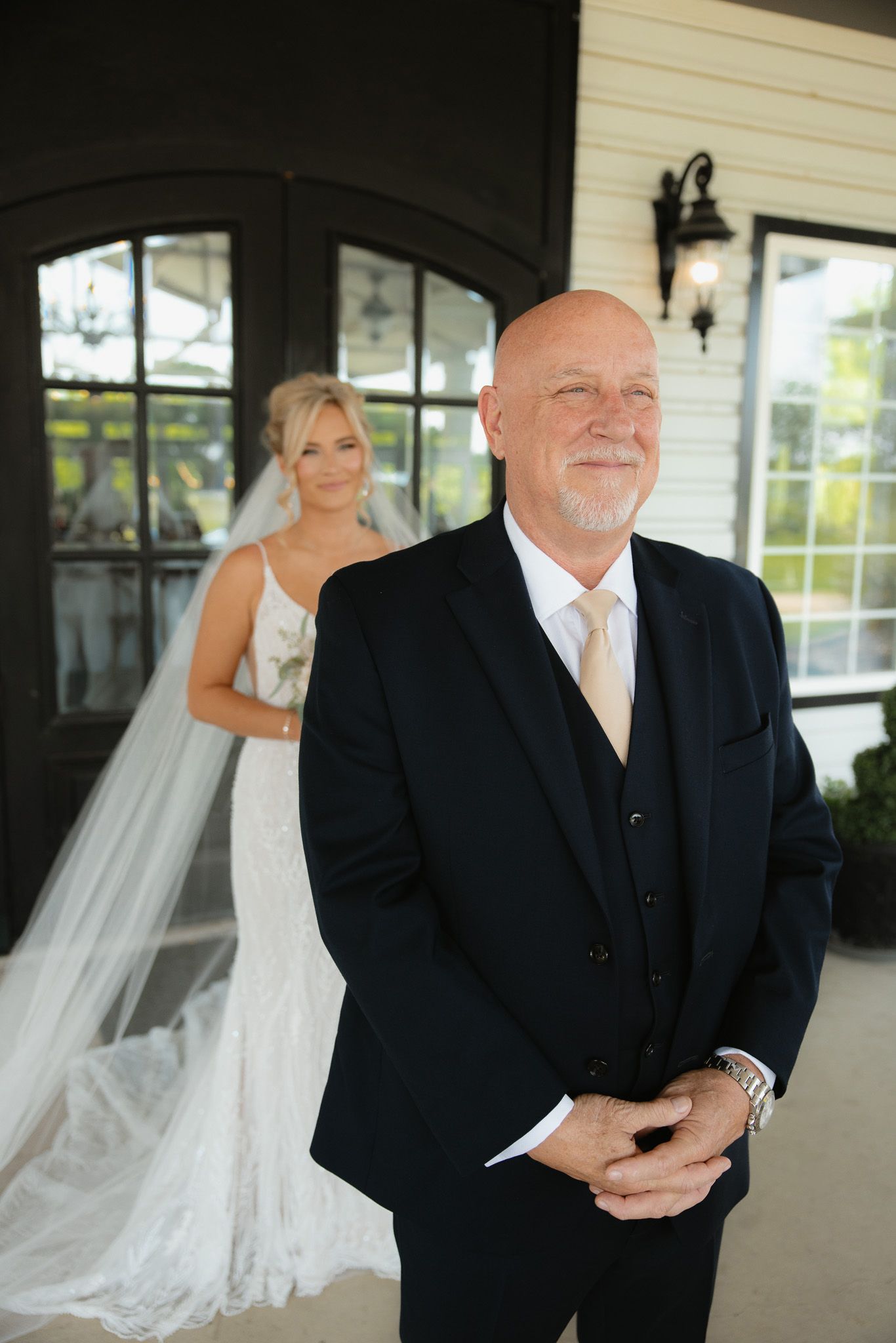 Bride in white wedding gown poses behind groom in black suit during first look moment at wedding venue.