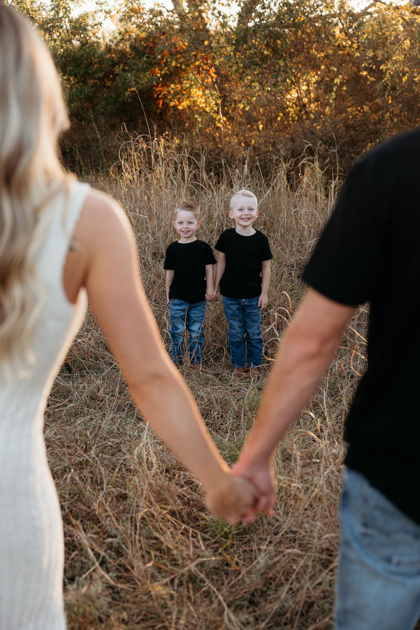 Couple holding hands in golden field at sunset with two children in matching outfits standing in background.