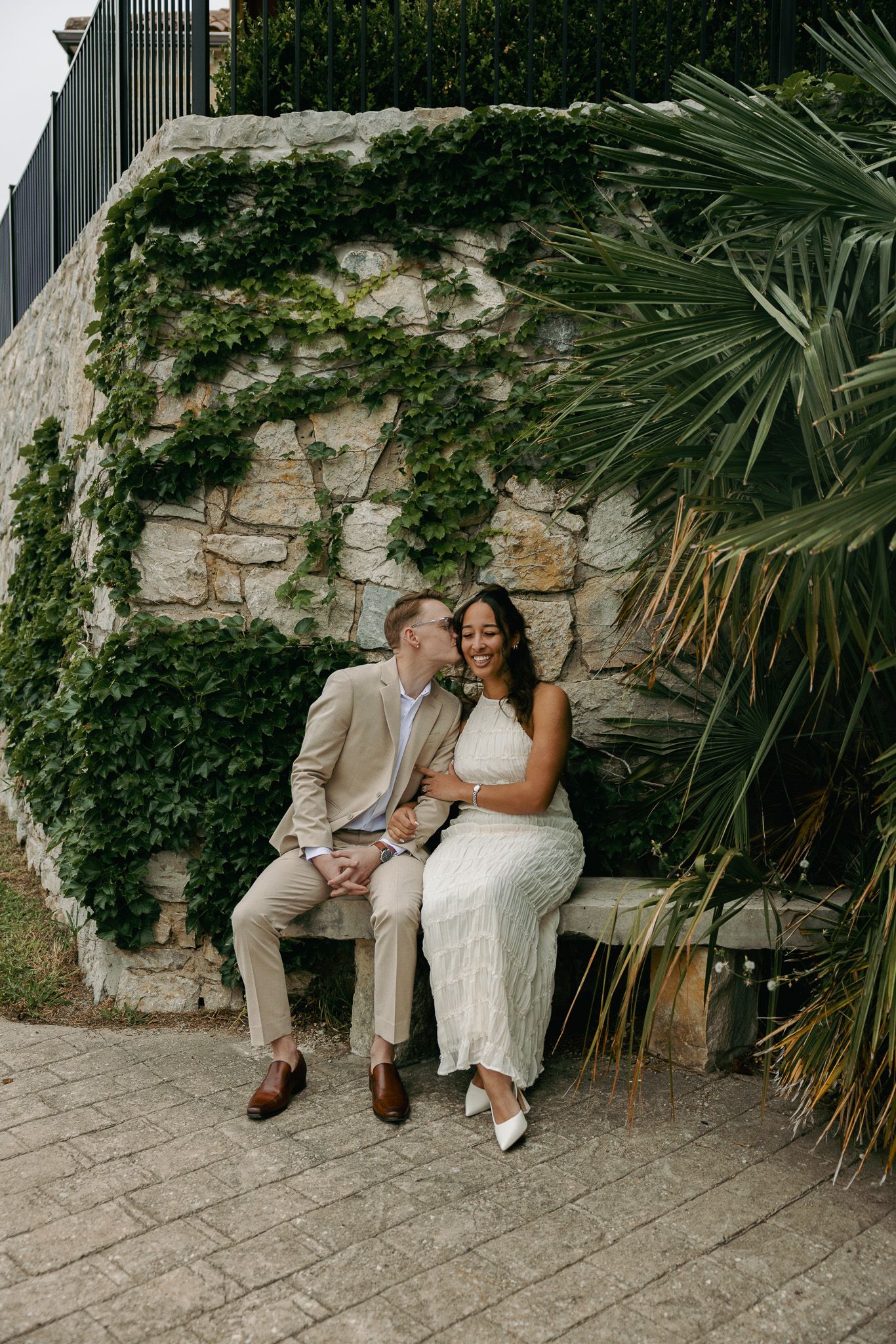 Couple in wedding attire sitting on a bench, groom kissing bride on cheek, ivy-covered stone wall backdrop.