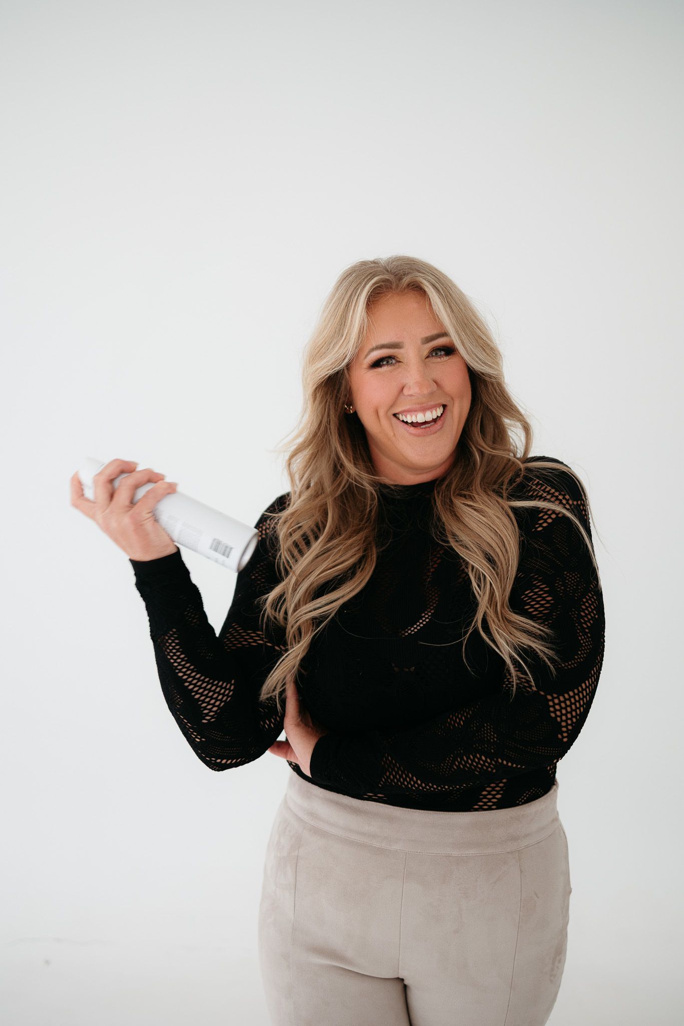 Smiling blonde woman in black mesh top and beige pants holding a white bottle against a white background.