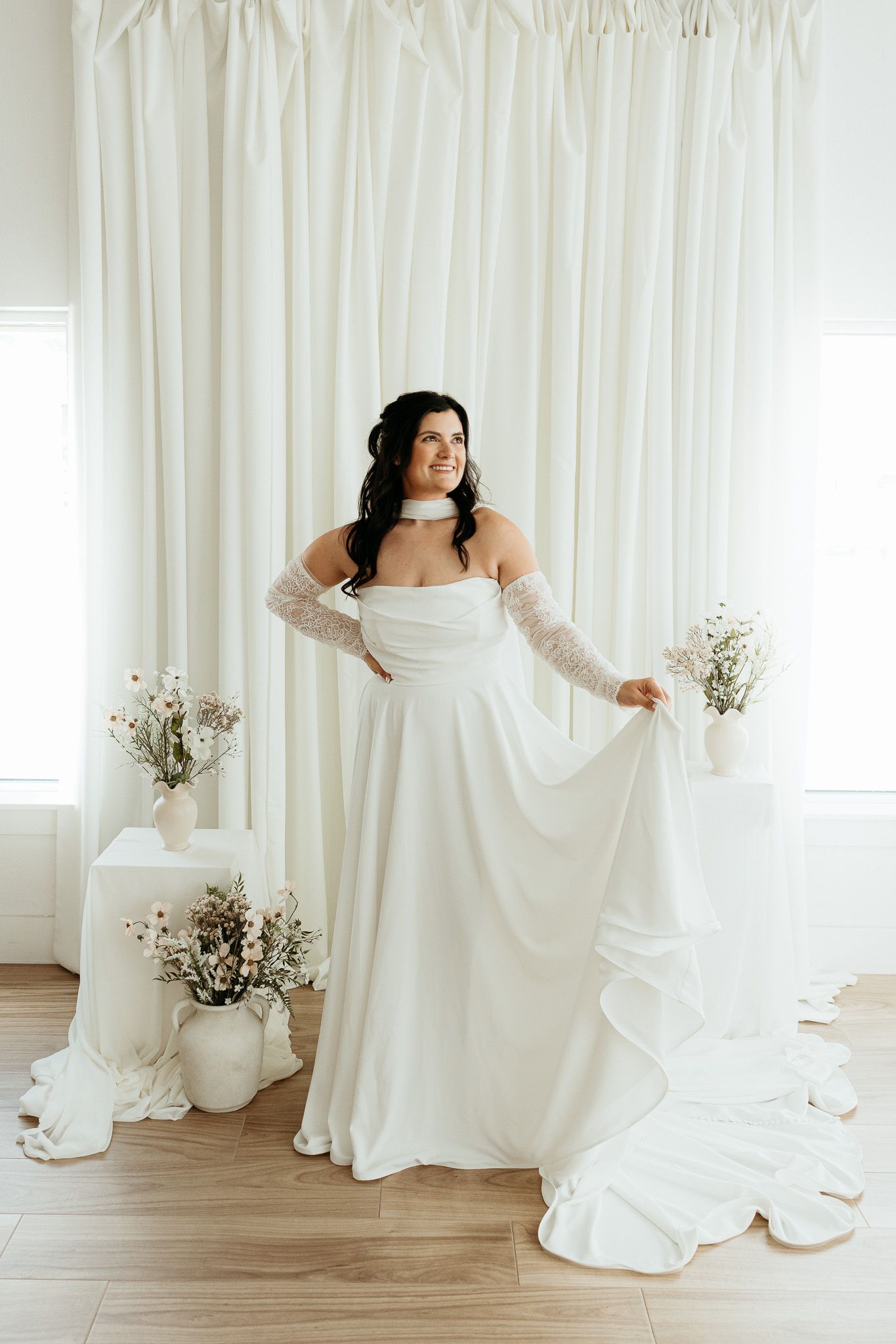 Bride in elegant off-shoulder white gown with lace gloves, holding dress train in bright studio with floral decor.
