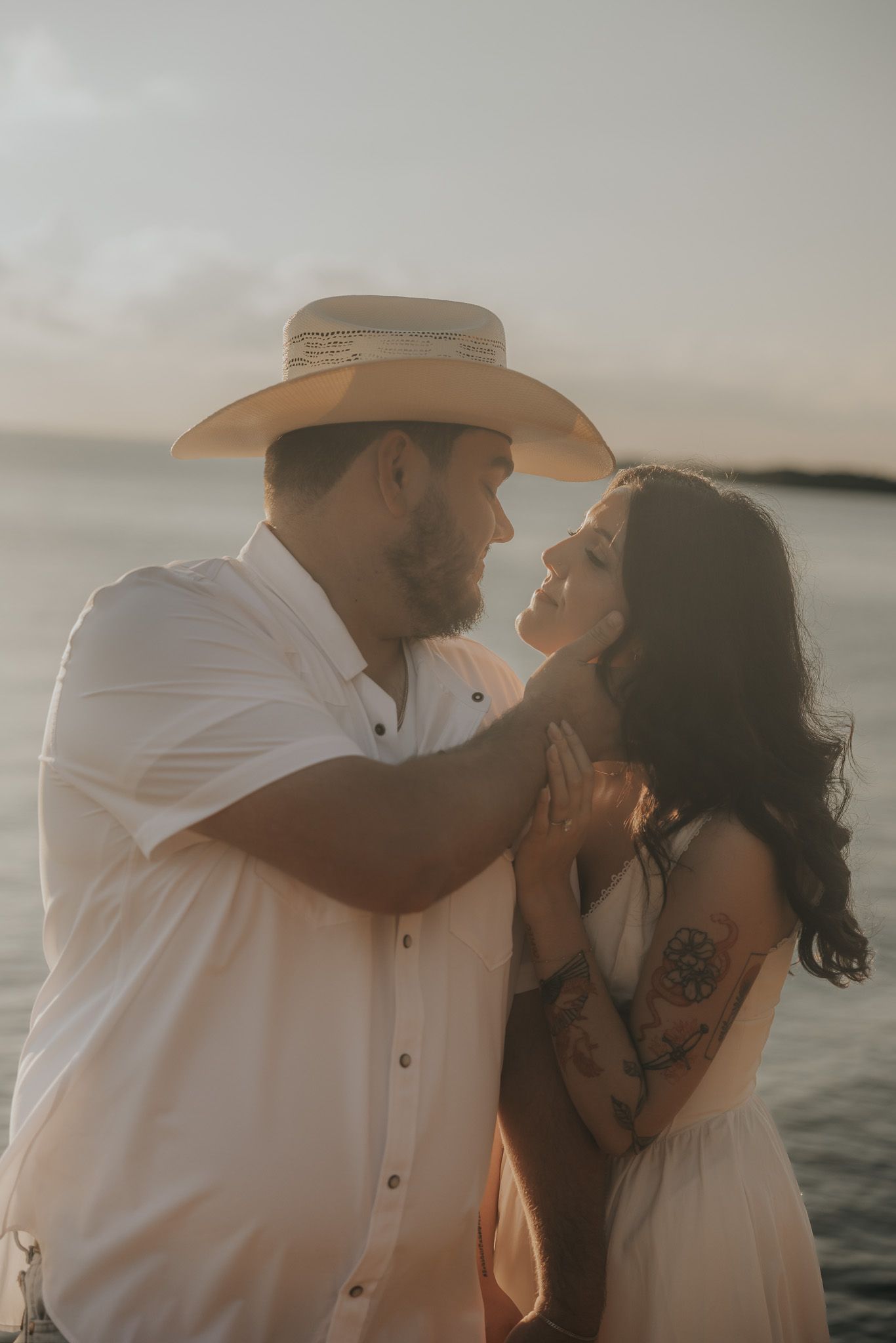 A romantic couple shares tender moments by the water during sunset, with the person wearing a cowboy hat and white shirt.
