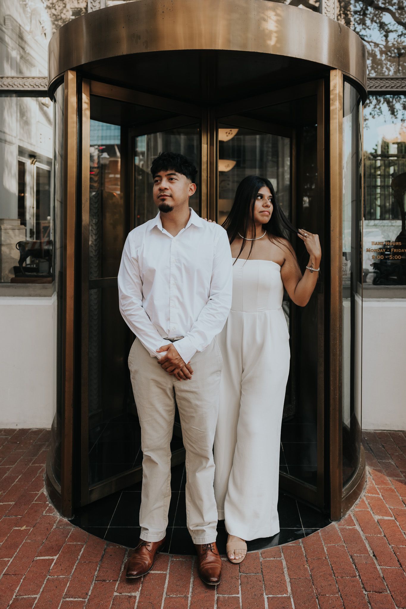 A couple in all-white outfits poses playfully near glass doors on a brick patio.