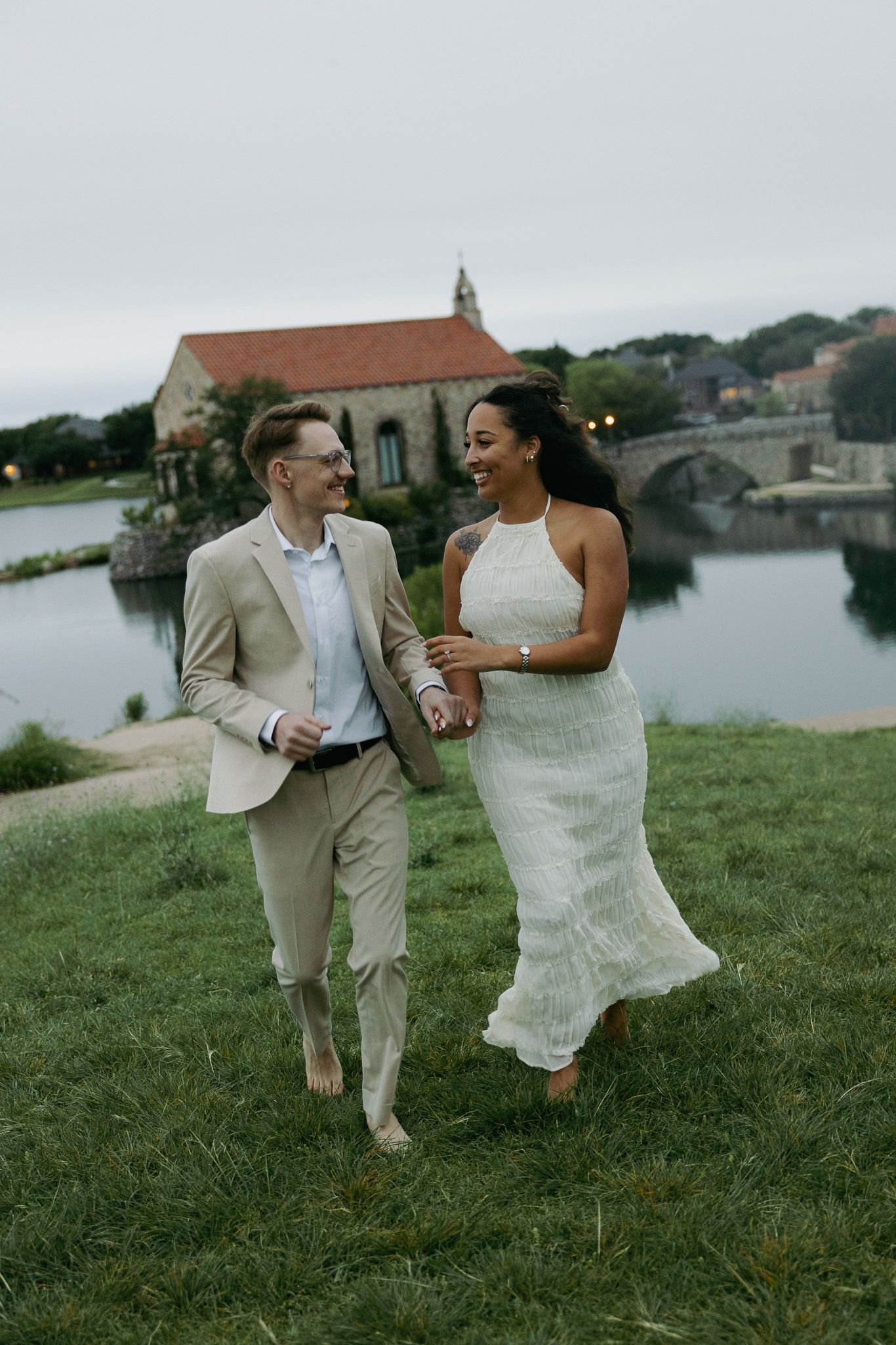 Couple in wedding attire walking barefoot on green grass by a waterfront at dusk, smiling at each other.