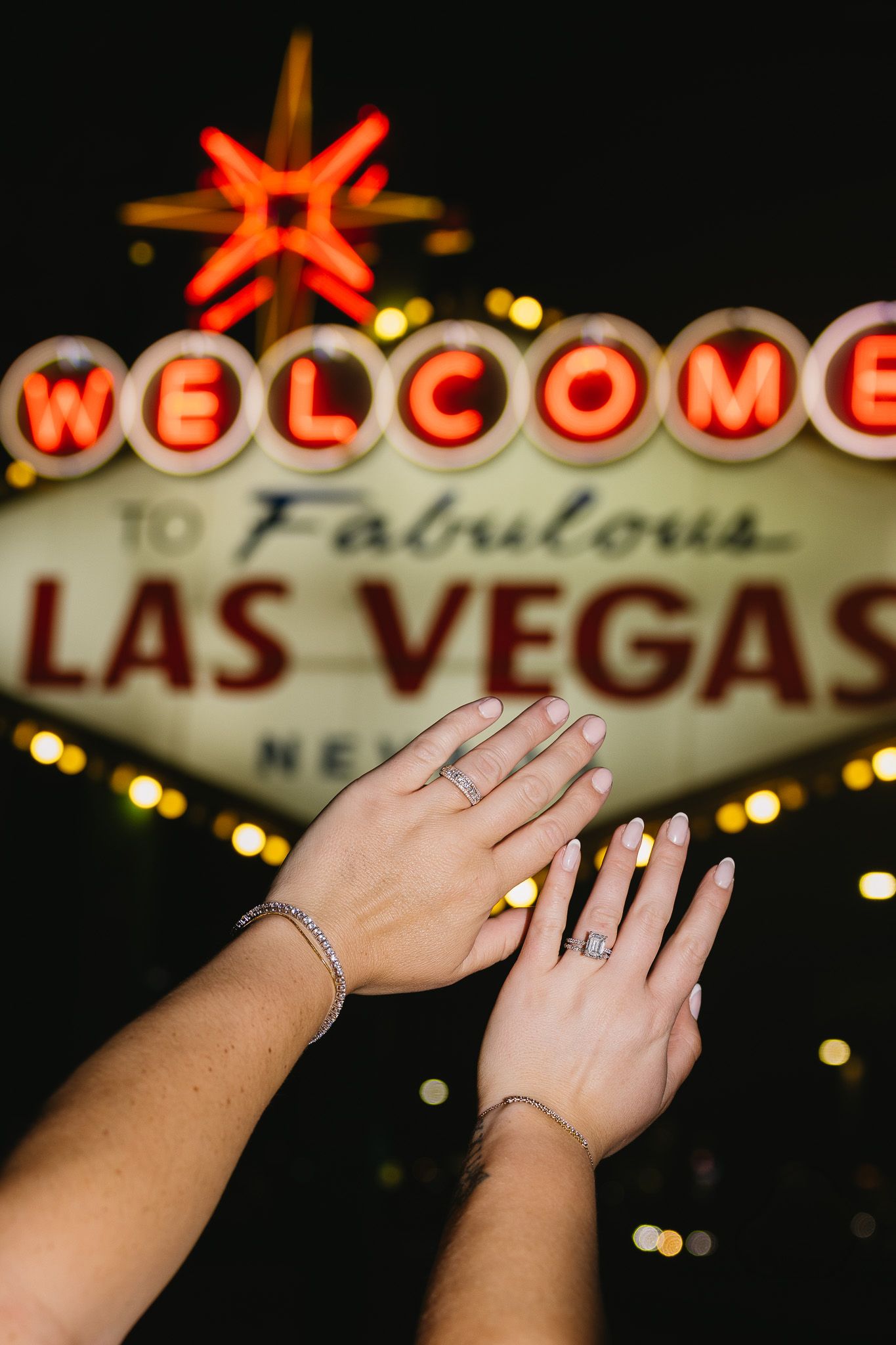 Two hands displaying engagement rings raised in front of the iconic Welcome to Fabulous Las Vegas neon sign at night.