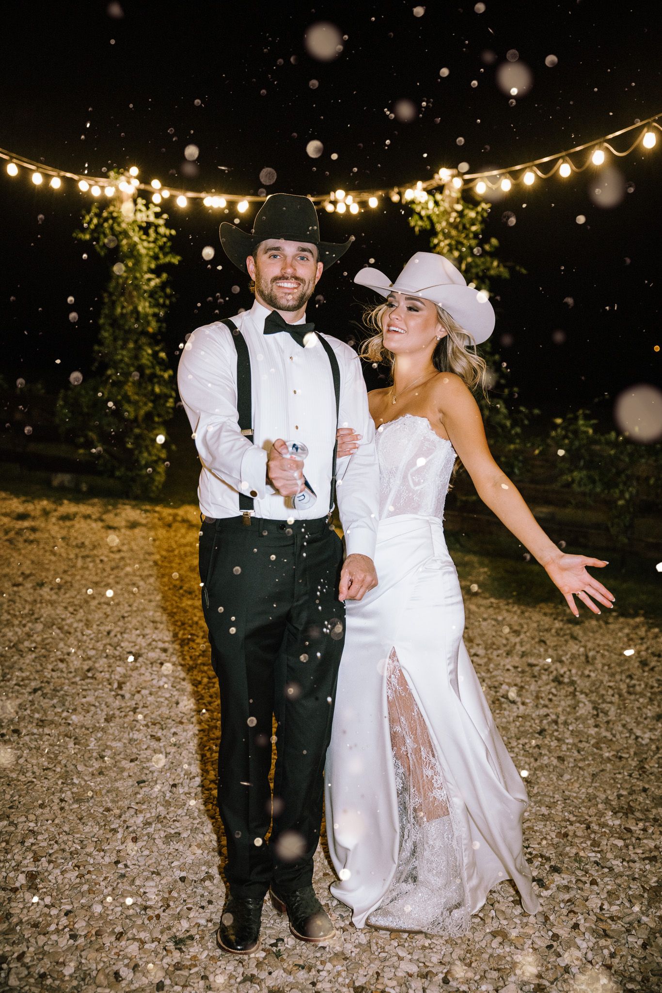 Bride and groom in cowboy hats celebrate outdoors at night with string lights and confetti.