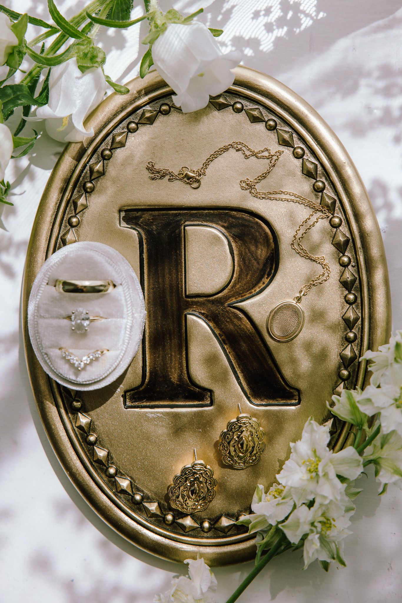 Gold oval monogram R plaque with wedding rings, necklace, and white flowers for elegant bridal jewelry flat lay.