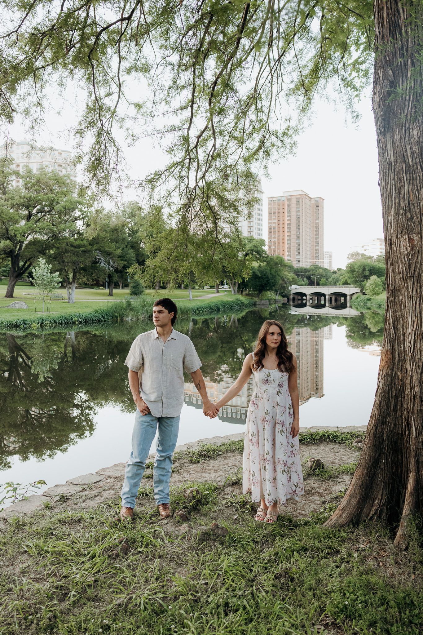 Couple holding hands by a serene park pond with city buildings reflected in the water beneath a large tree.
