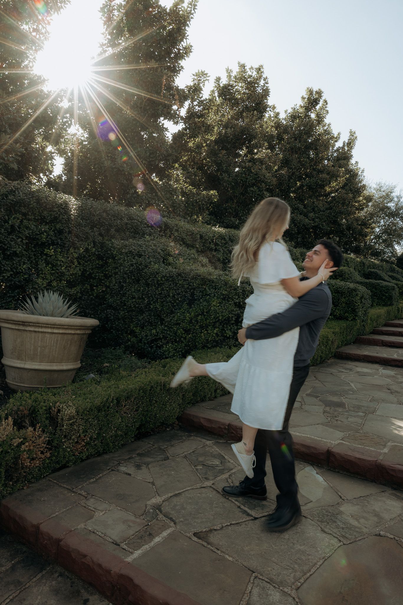 A couple dances together on a stone patio at sunset with trees in the background.