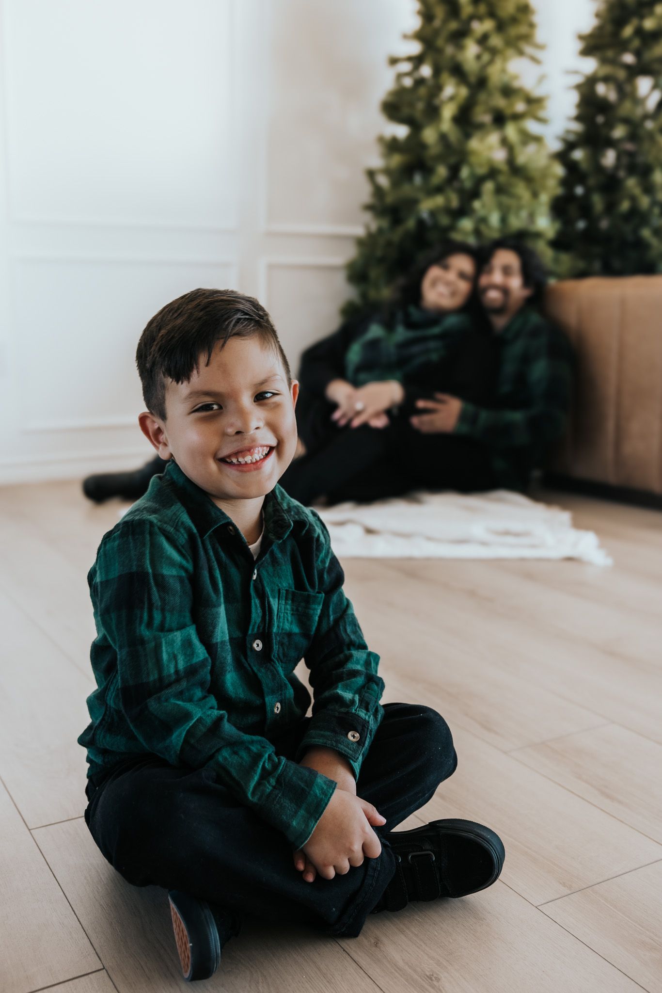A child in a green plaid shirt sits cross-legged on a wooden floor with a bright smile.