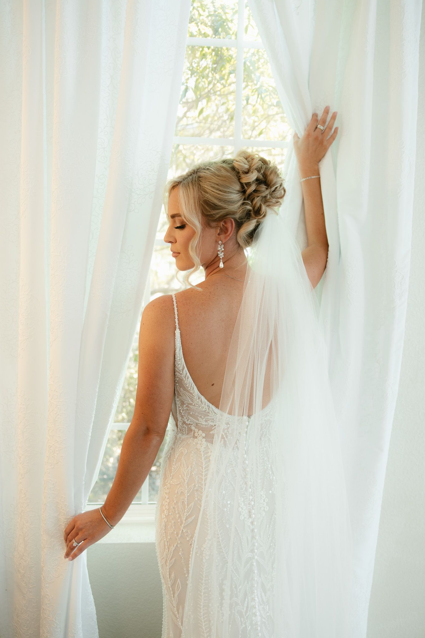 Elegant bride in a backless lace wedding gown with veil and updo, standing by sheer white curtains near a window.