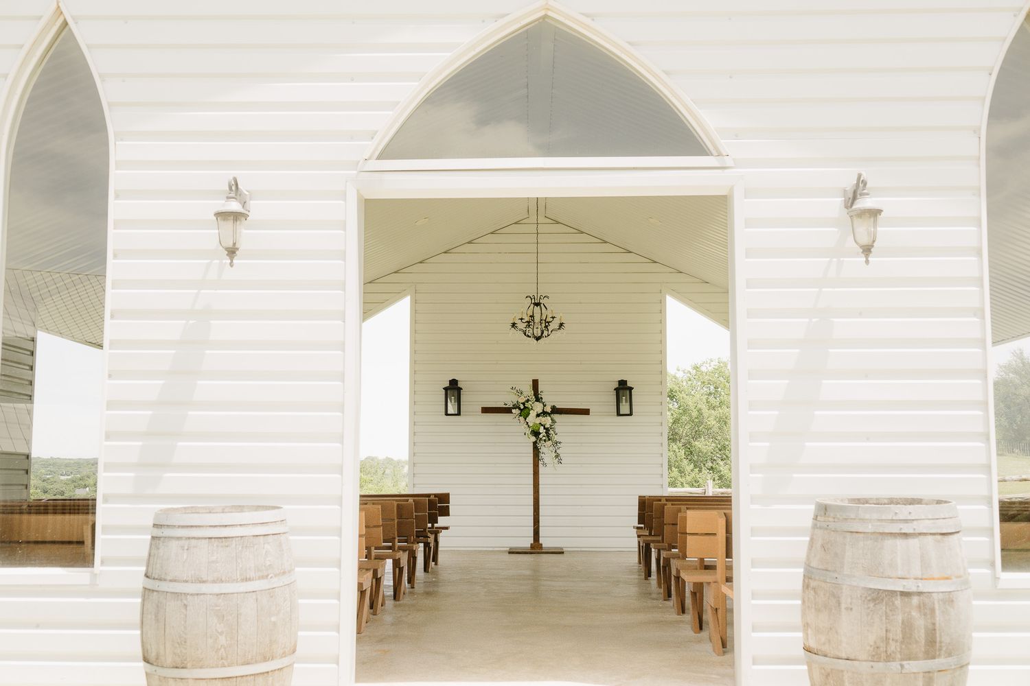 White chapel interior with wooden pews, floral cross altar, chandelier, and barrel decor at a rustic wedding venue.