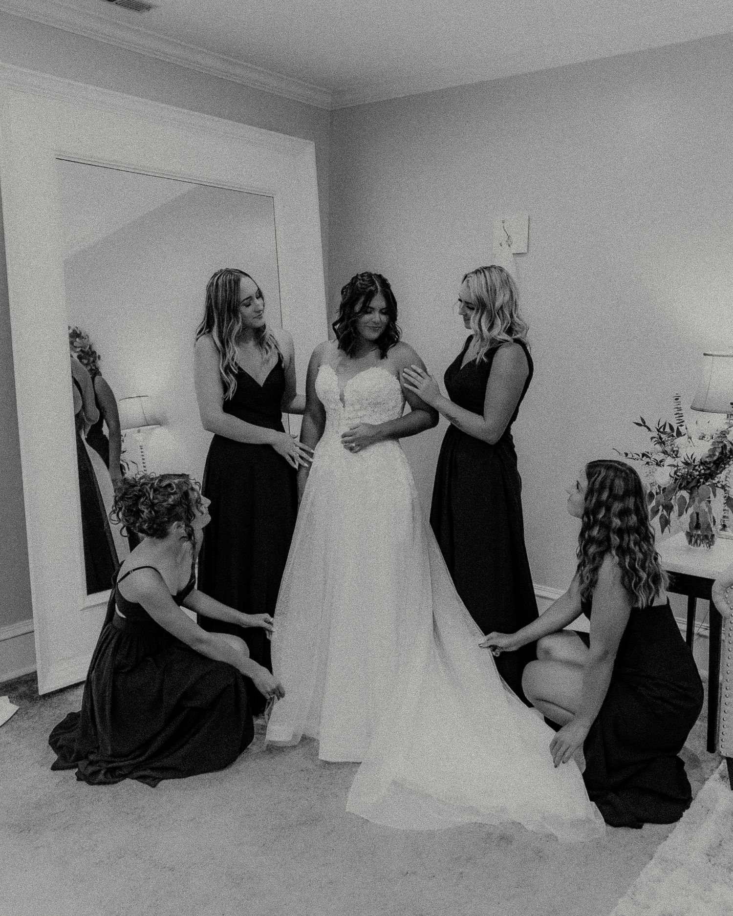Bridesmaids in black dresses pose with a wedding gown in a getting-ready room.