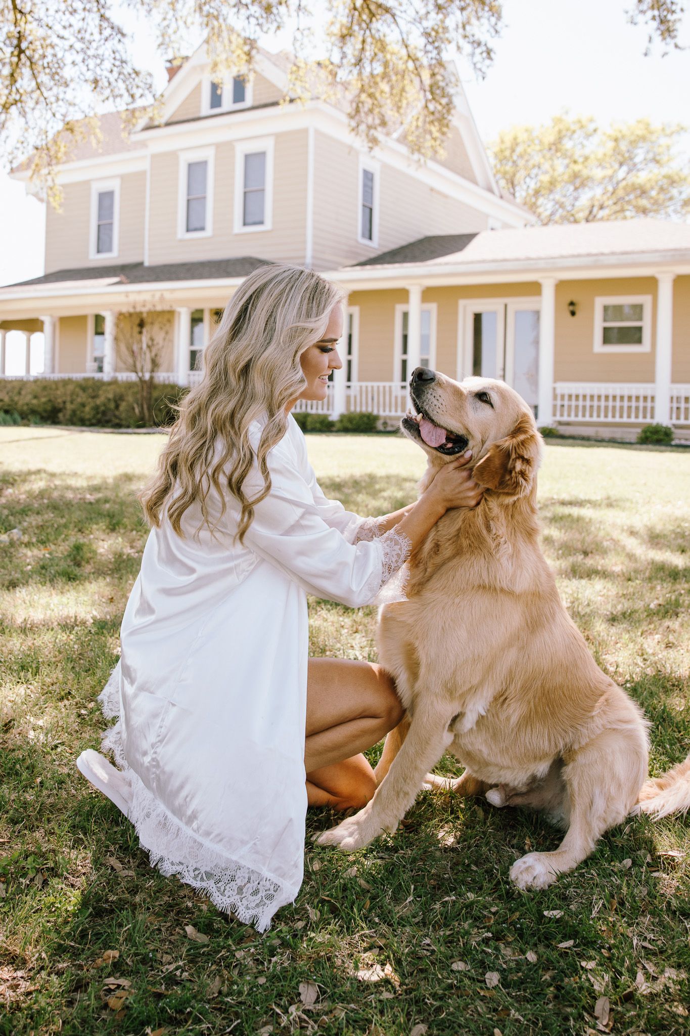 Woman in white dress petting a golden retriever on green lawn in front of a yellow farmhouse.