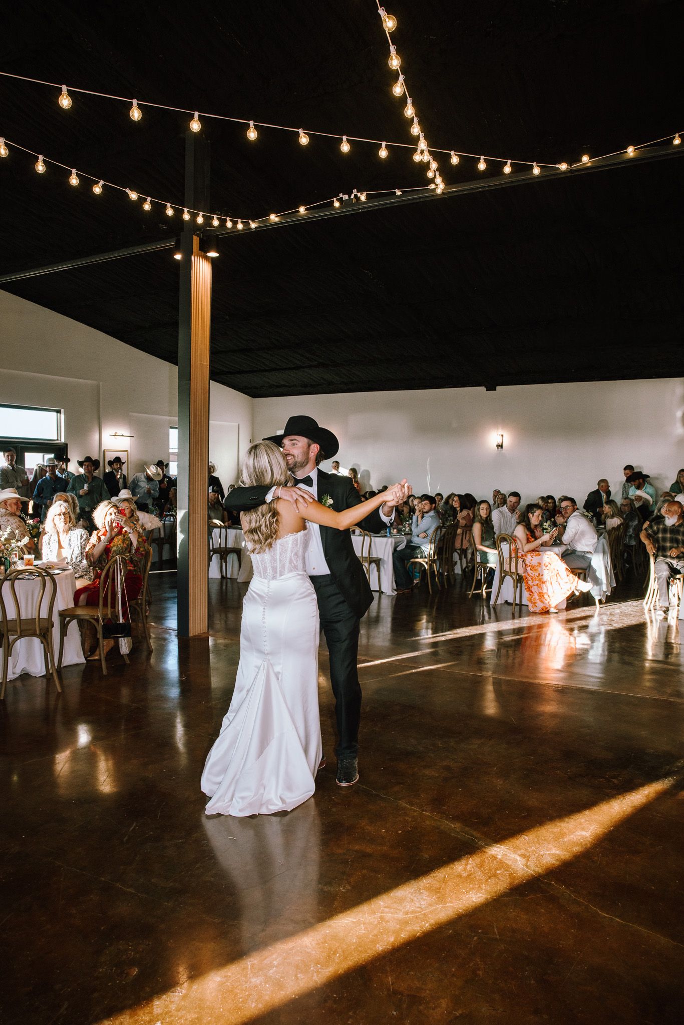 Bride and groom share first dance under string lights at rustic wedding venue with guests seated in background.