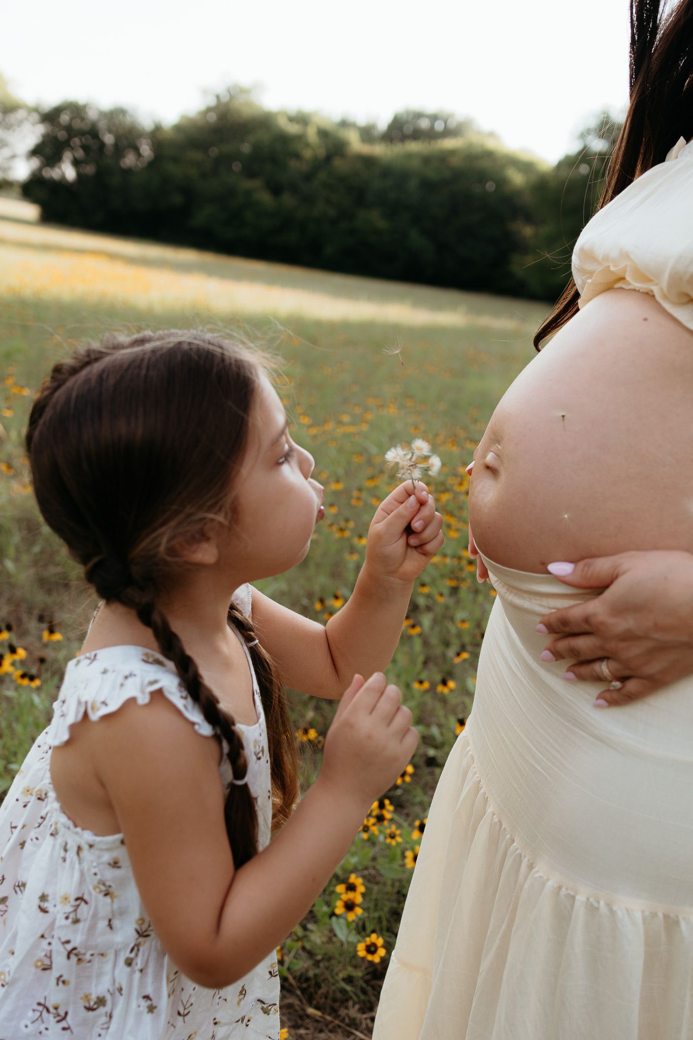 A young girl in a floral dress blows a dandelion at a pregnant woman's belly in a sunny wildflower field.